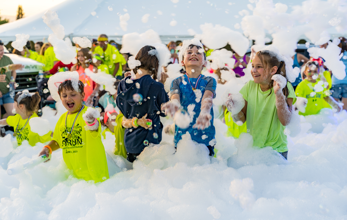 Children playing in foam at outdoor event during daytime, with some wearing bright yellow shirts, smiling and laughing.