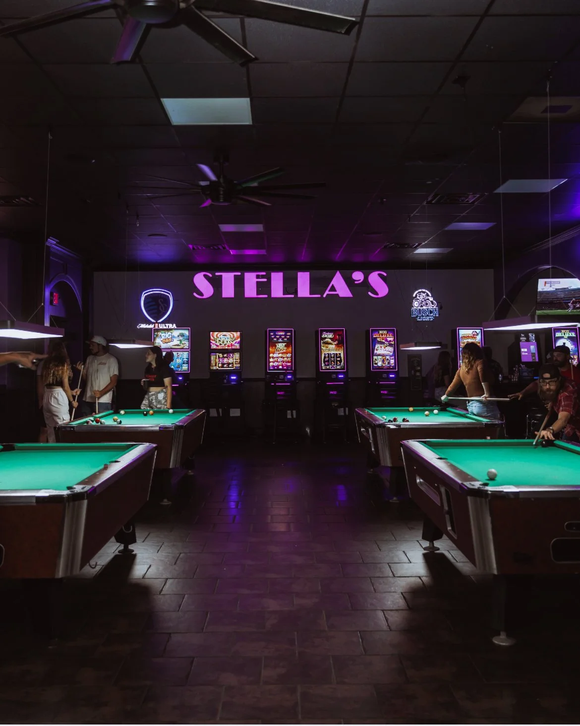 An indoor billiard hall called Stella's with pool tables, neon signs, and people playing or standing around.