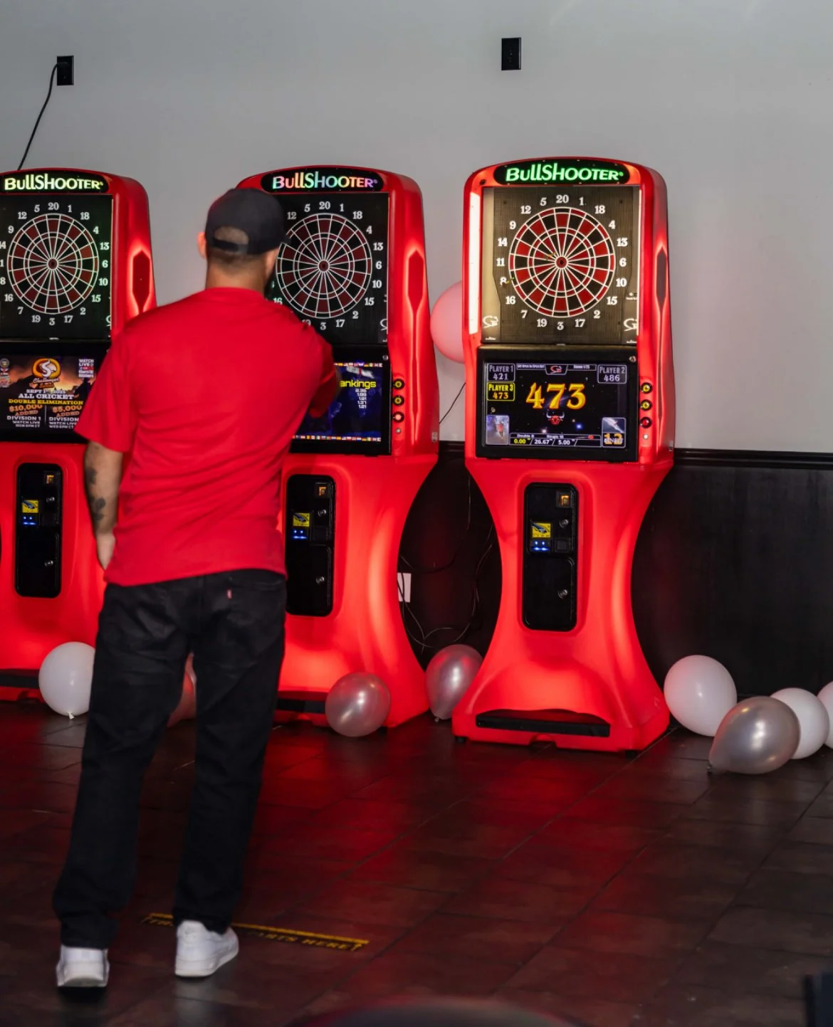 A man in a red shirt and black pants playing darts on an electronic dartboard in a decorated room.