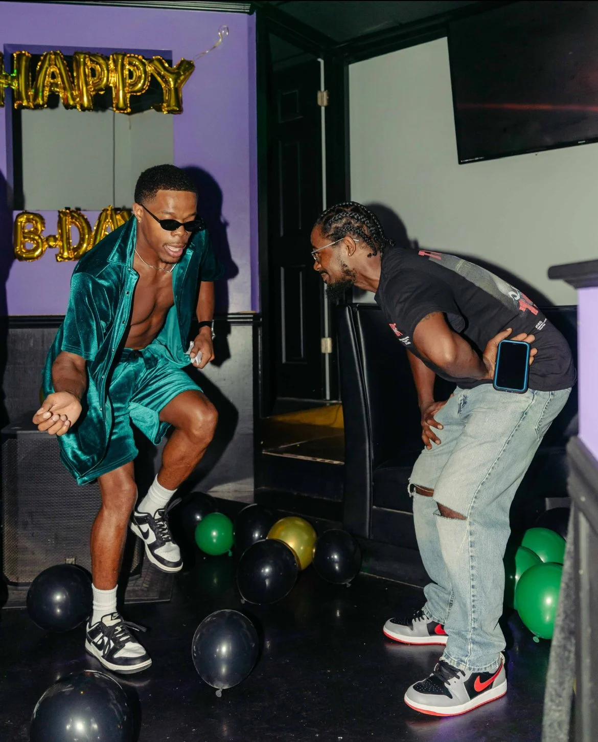 Two men are celebrating at a birthday party with balloons and a 'Happy B-Day' banner in the background. One man is wearing sunglasses, a teal satin shirt and shorts, and sneakers, while the other man is bending over, smiling, holding a phone, and dressed in casual ripped jeans and sneakers.