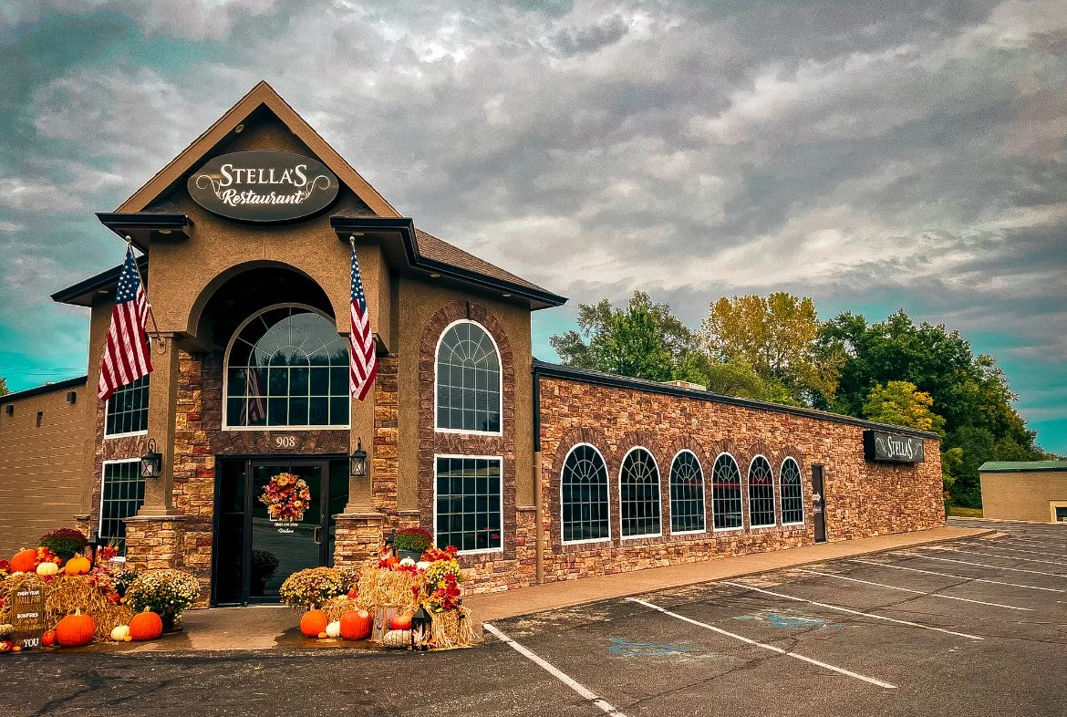 Exterior of Stella's Restaurant with fall decorations, pumpkins, and flowers in front, American flags, and a stone and stucco facade, under a cloudy sky.