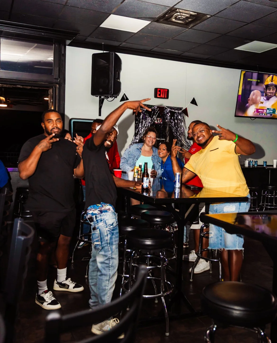 A group of friends at a social gathering in a bar or restaurant, posing for a photo around a high table with drinks, some making peace signs and smiling happily.