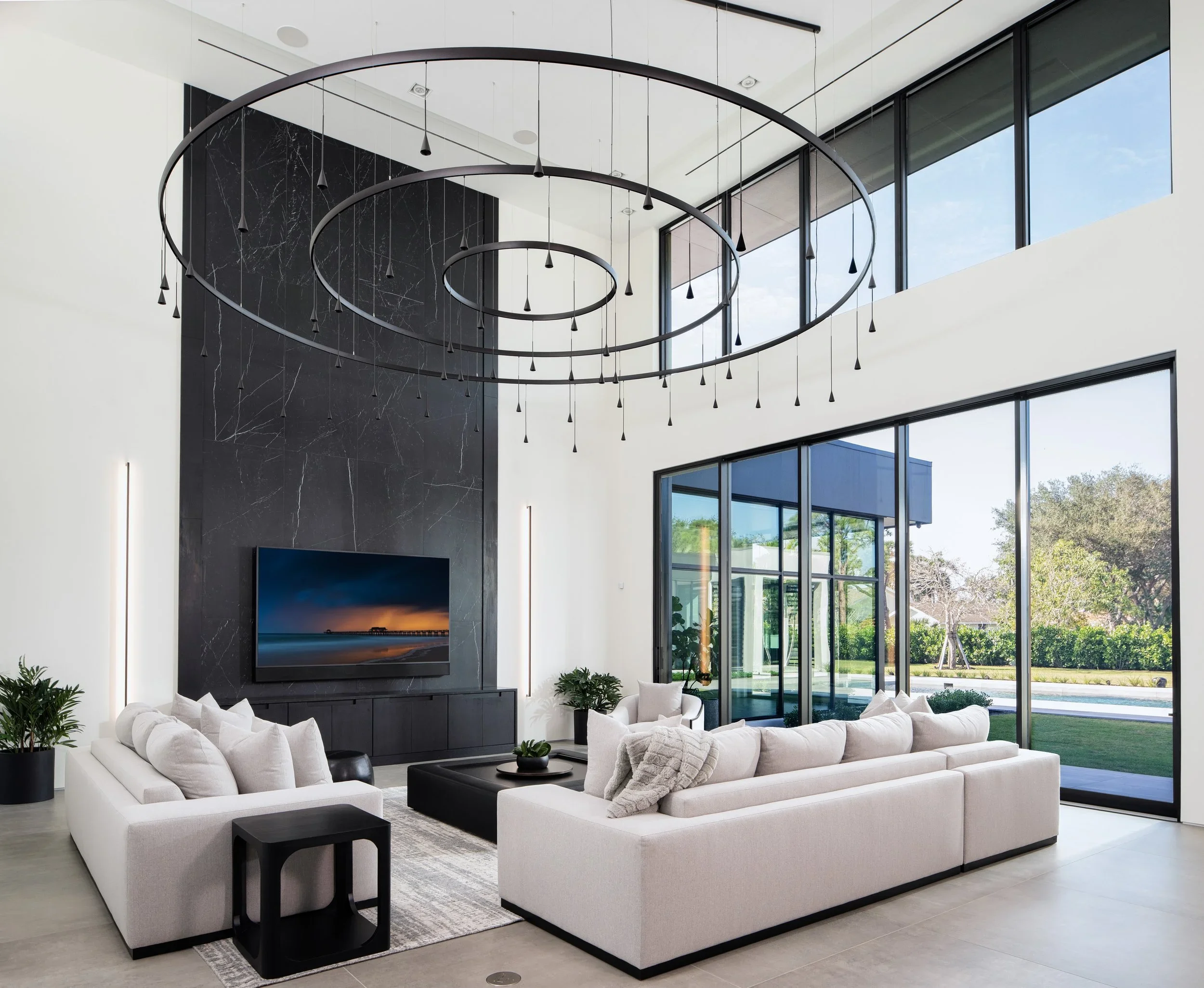 Modern living room with large windows, white sofas, black coffee table, and a flat-screen TV on a black marble accent wall, with a spiral chandelier hanging from the ceiling.