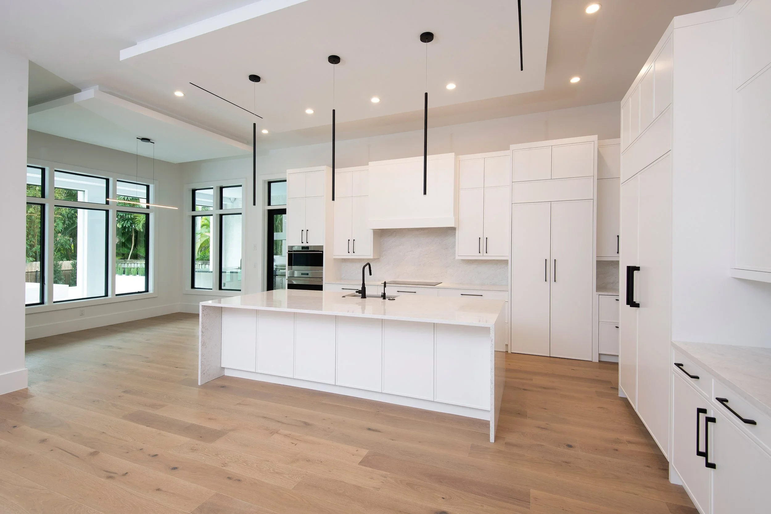 Modern white kitchen with black handles, light wood flooring, large windows, and a kitchen island with black sink and fixtures. Ceiling lights and a minimalist design.