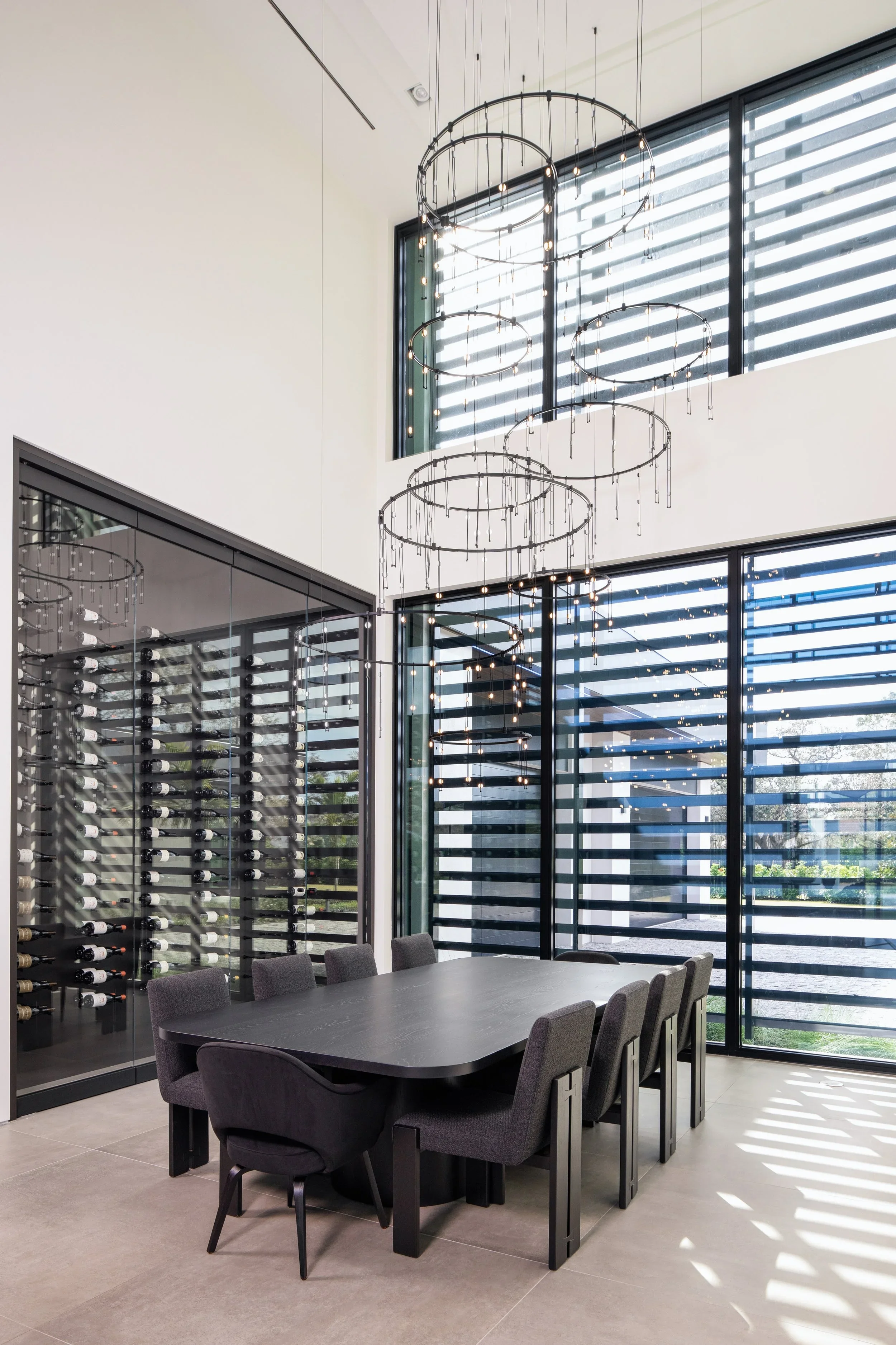 Modern dining room with a black table and six gray chairs, floor-to-ceiling windows with horizontal shades, a wine storage unit, and a contemporary chandelier with multiple circular metal rings and small lights.