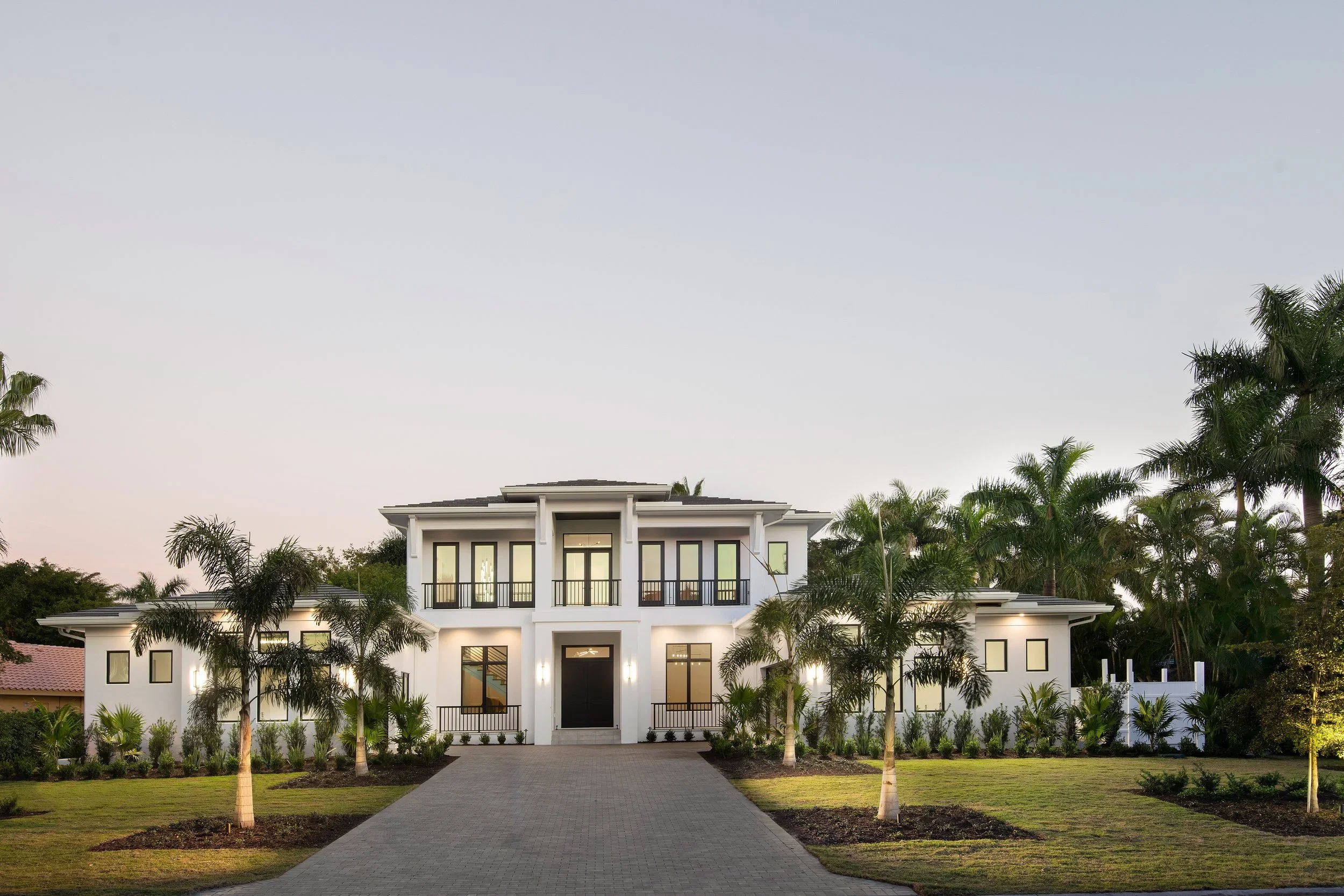 Large modern white house with multiple balconies, surrounded by lush palm trees and a well-maintained lawn, under a clear sky.