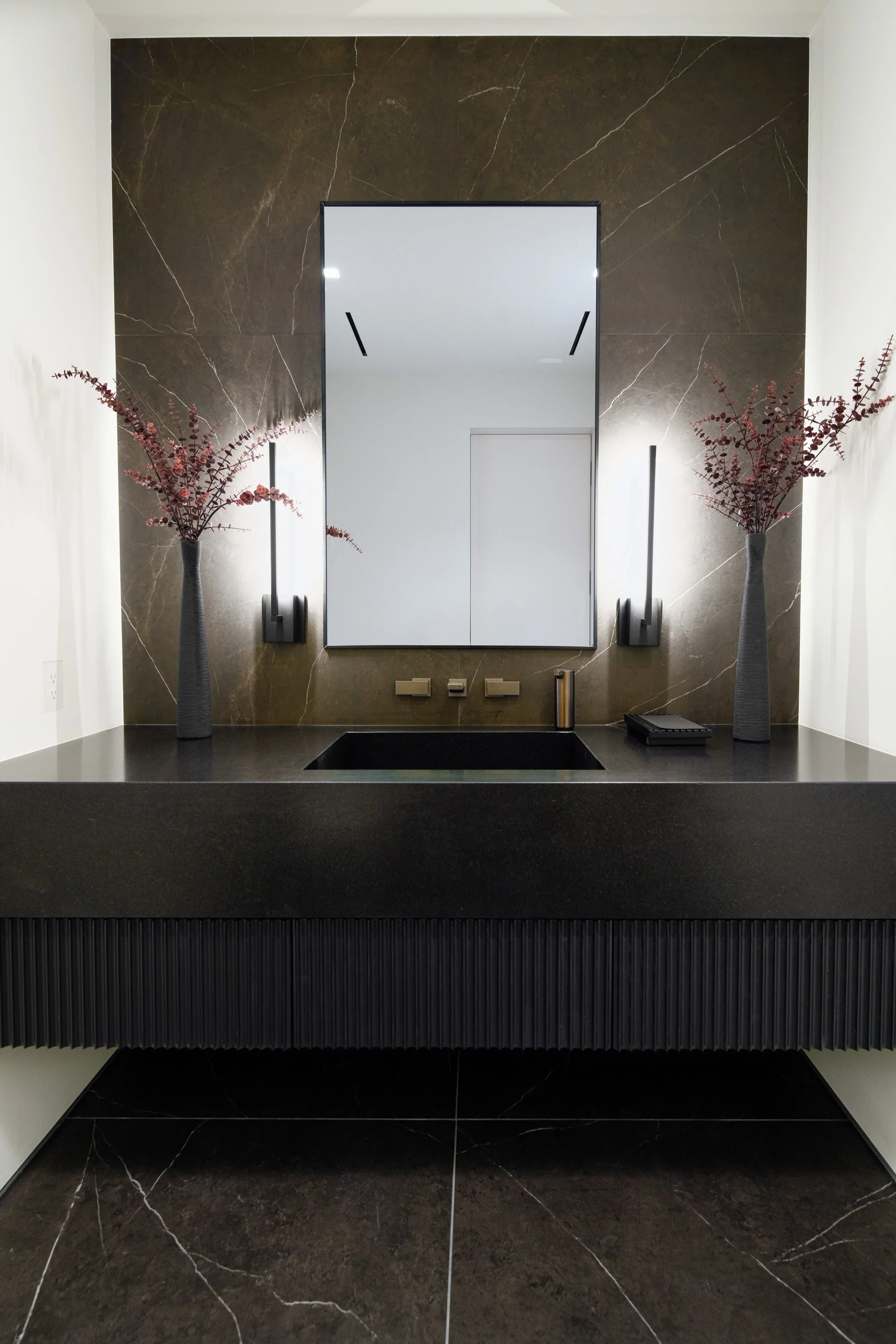 Modern bathroom vanity with a black countertop, a large mirror, two tall flower vases with red branches, wall-mounted black light fixtures, and a dark marble wall background.