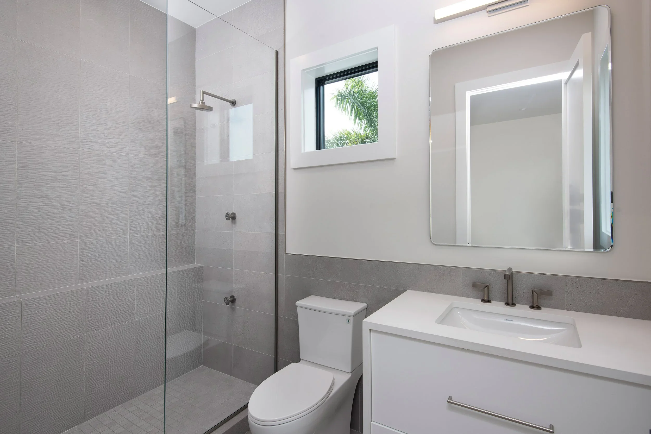 A modern bathroom with a walk-in shower, a white toilet, a white vanity with a sink, a large mirror, and a window showing green foliage outside.