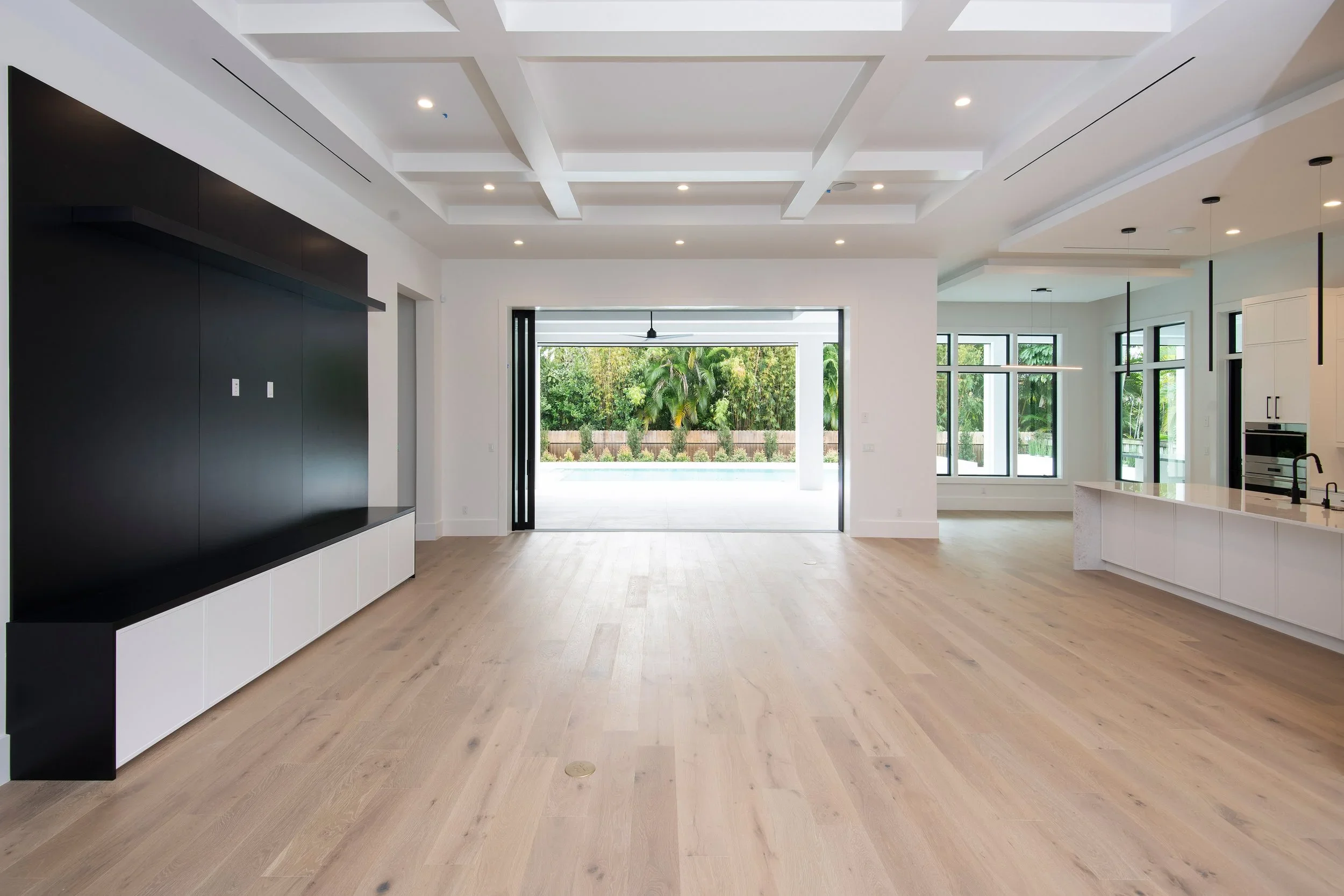 Interior of a modern, spacious living area with open floor plan, large windows, light wood flooring, and a view of a backyard with greenery and pool.