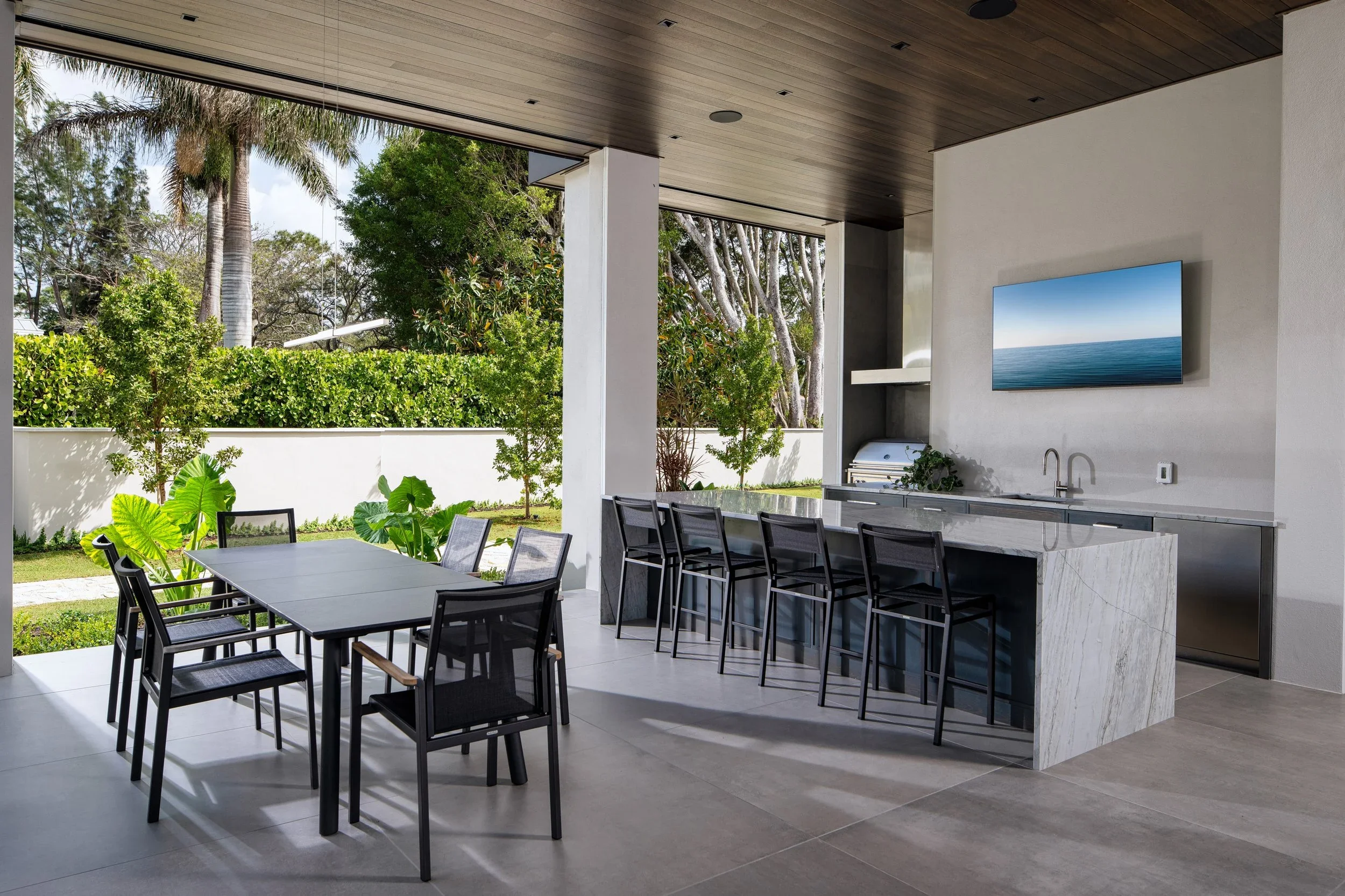 Modern outdoor kitchen and dining area with a black dining table and six chairs, a marble kitchen island with five bar stools, a wall-mounted TV displaying an ocean scene, a barbecue grill, and lush green trees outside.