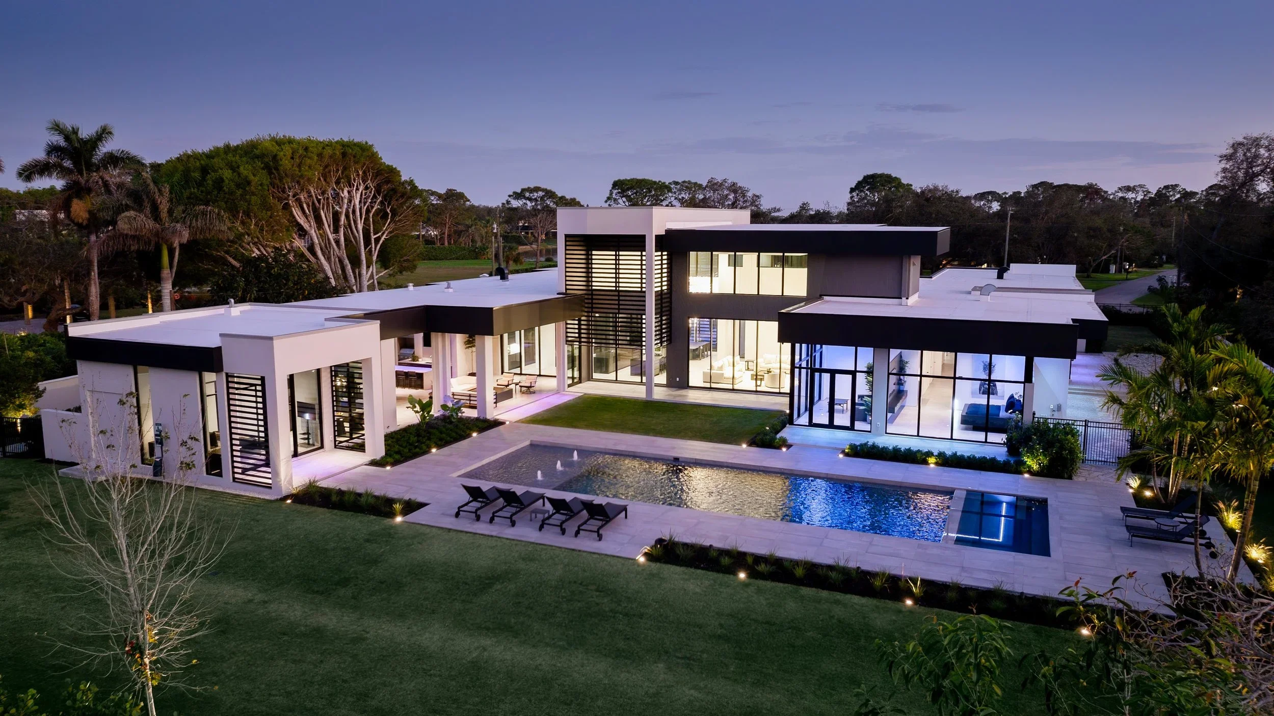 Modern house with large windows, outdoor pool, and landscaped yard at dusk, surrounded by trees and streetlights.