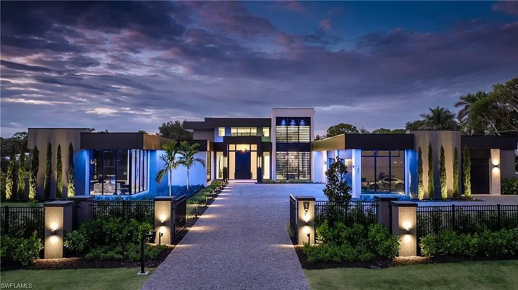 Modern luxury house with exterior lighting, a driveway, palm trees, and landscaped yard during dusk.