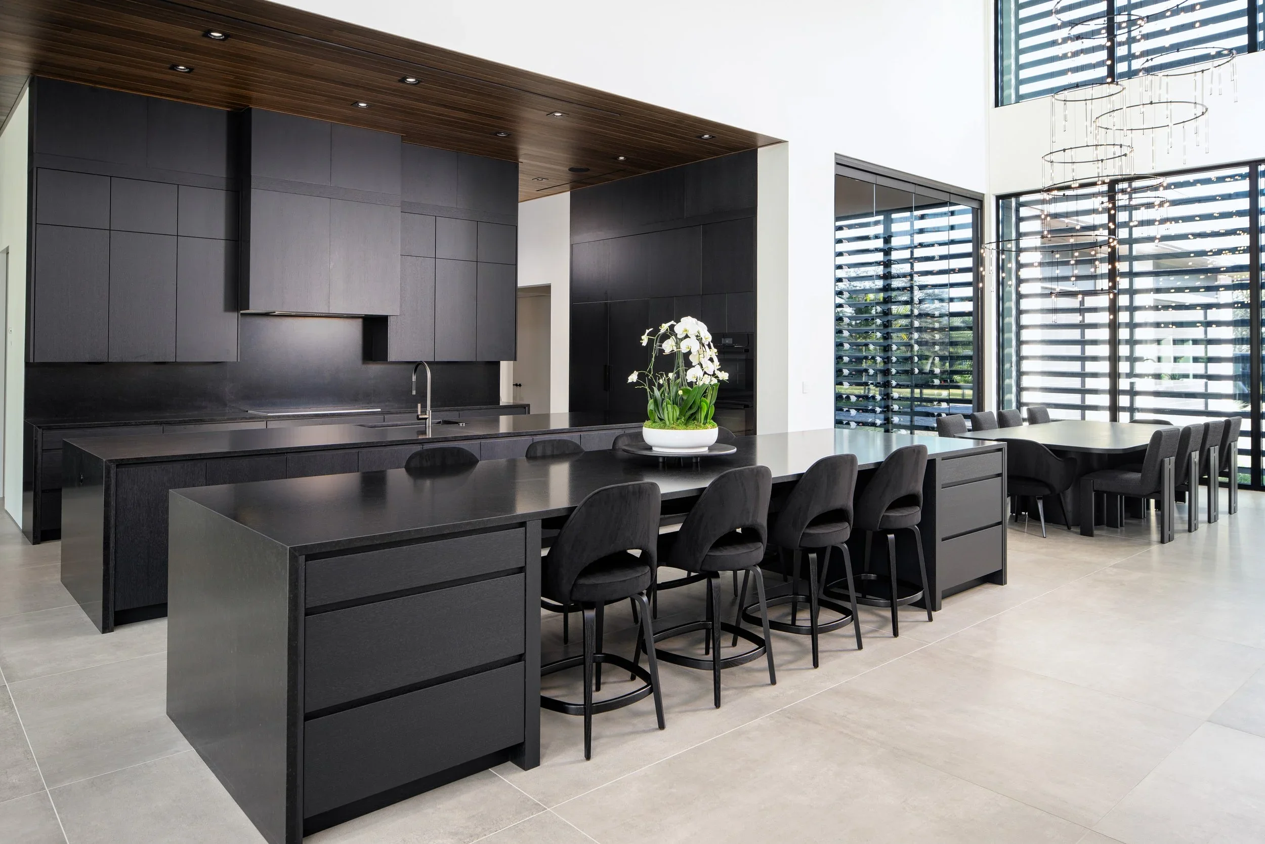 Modern kitchen with black cabinets, a black island, white walls, a wooden ceiling, a large window with black horizontal blinds, and a dining table with chairs, featuring a white orchid plant in the center.