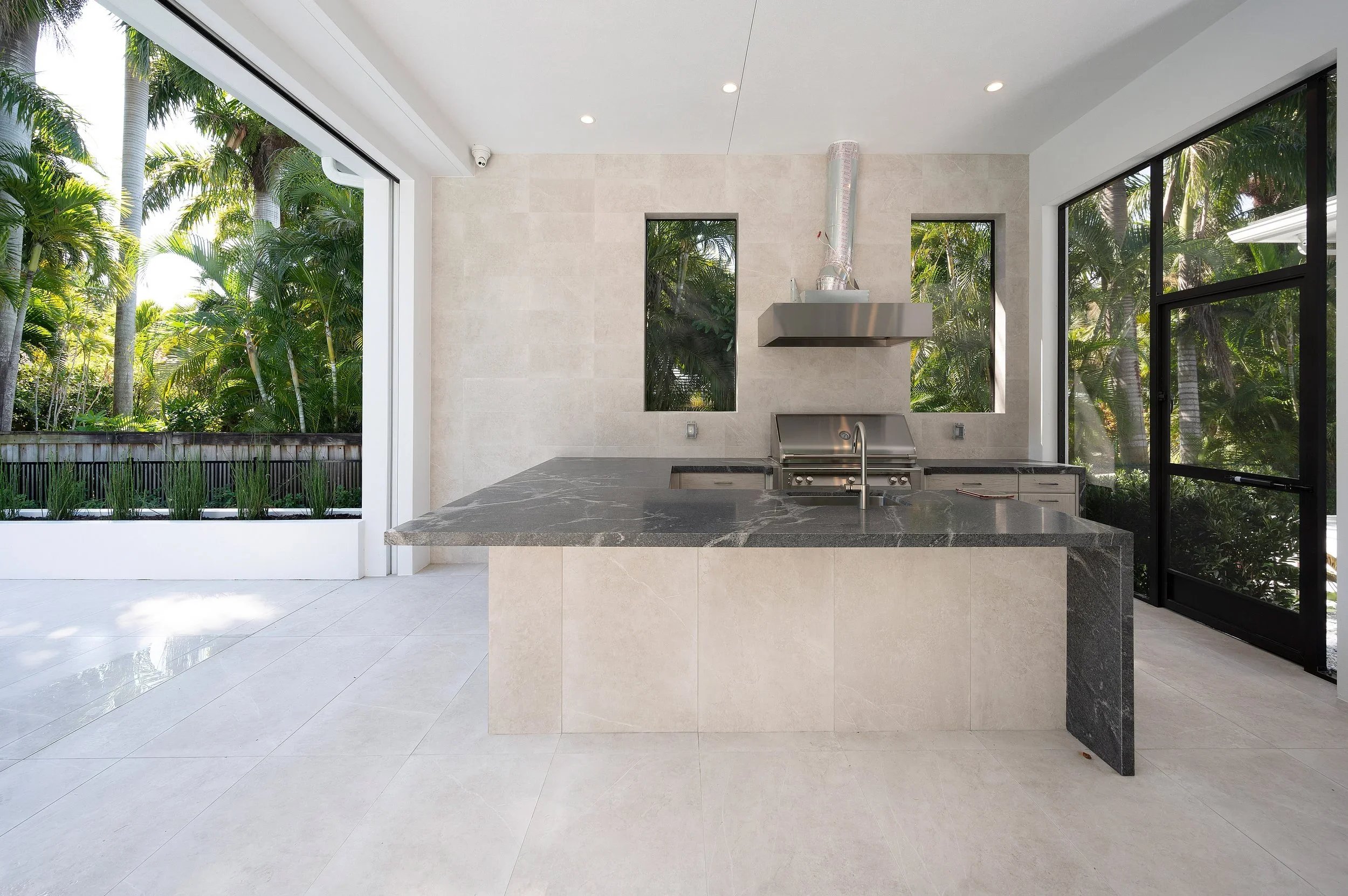 Modern outdoor kitchen with a black marble island, stainless steel grill, and large windows showing tropical greenery.