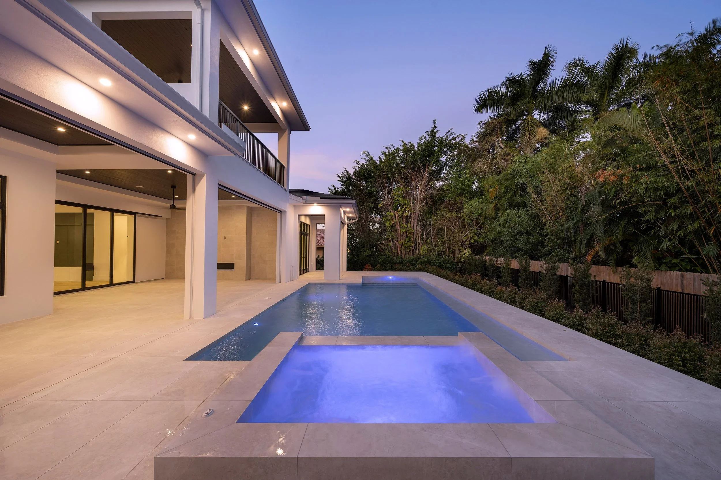 Modern backyard with swimming pool and hot tub, part of a house with large windows and upper-level balcony, surrounded by lush trees during dusk.