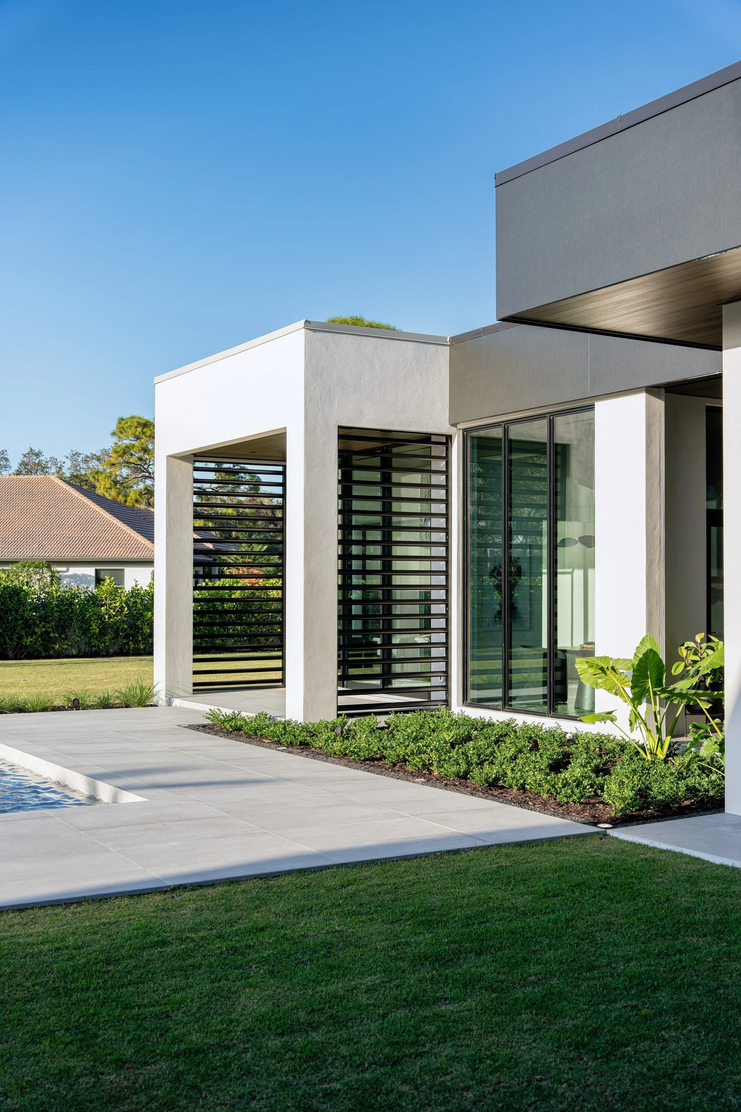 Modern house with large glass sliding doors, gray and white exterior walls, black horizontal slats, and landscaped yard with green grass and plants.