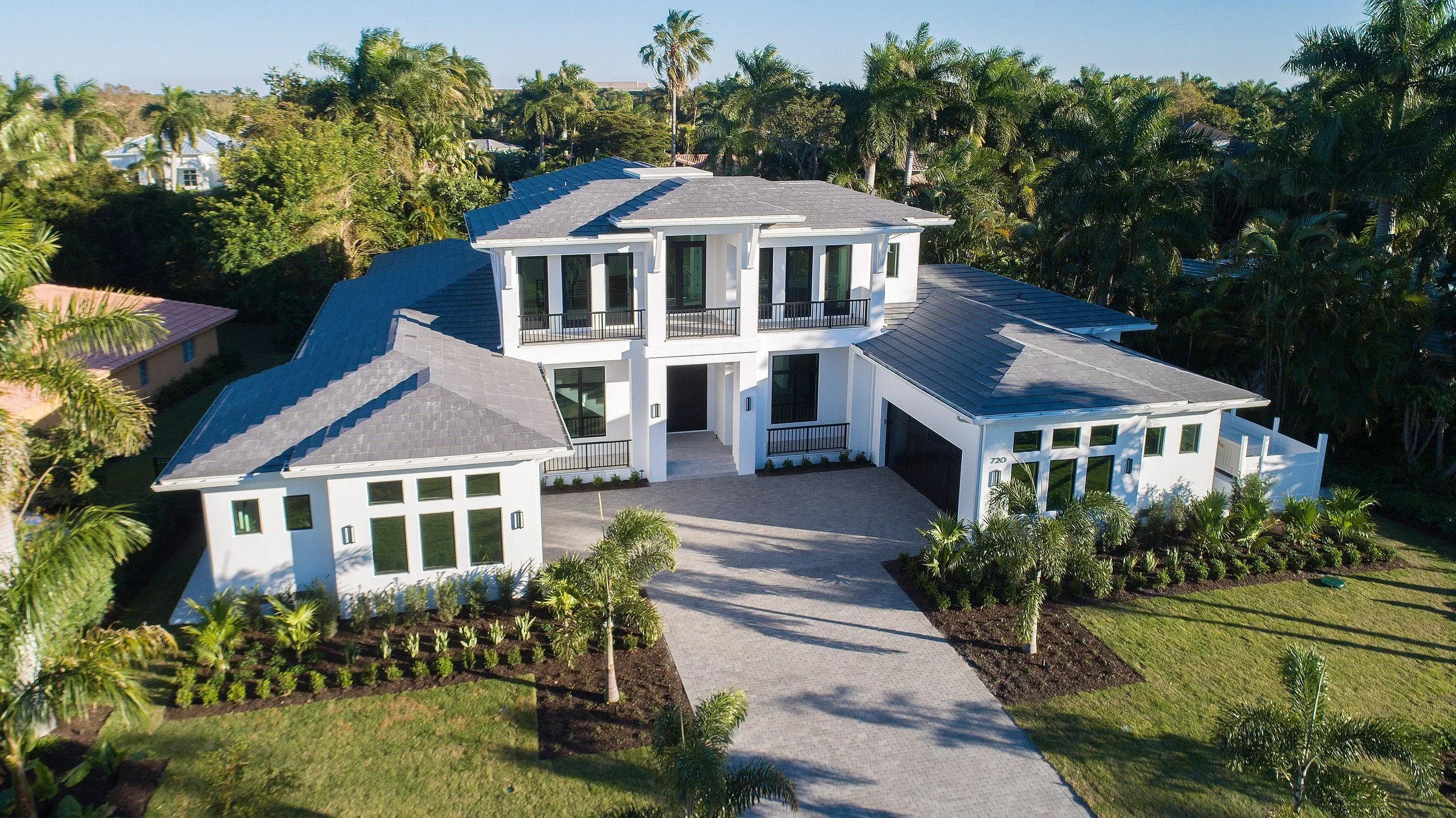 A modern two-story white house with a gray tiled roof, surrounded by palm trees and landscaped lawn, with a paved driveway leading to the garage.