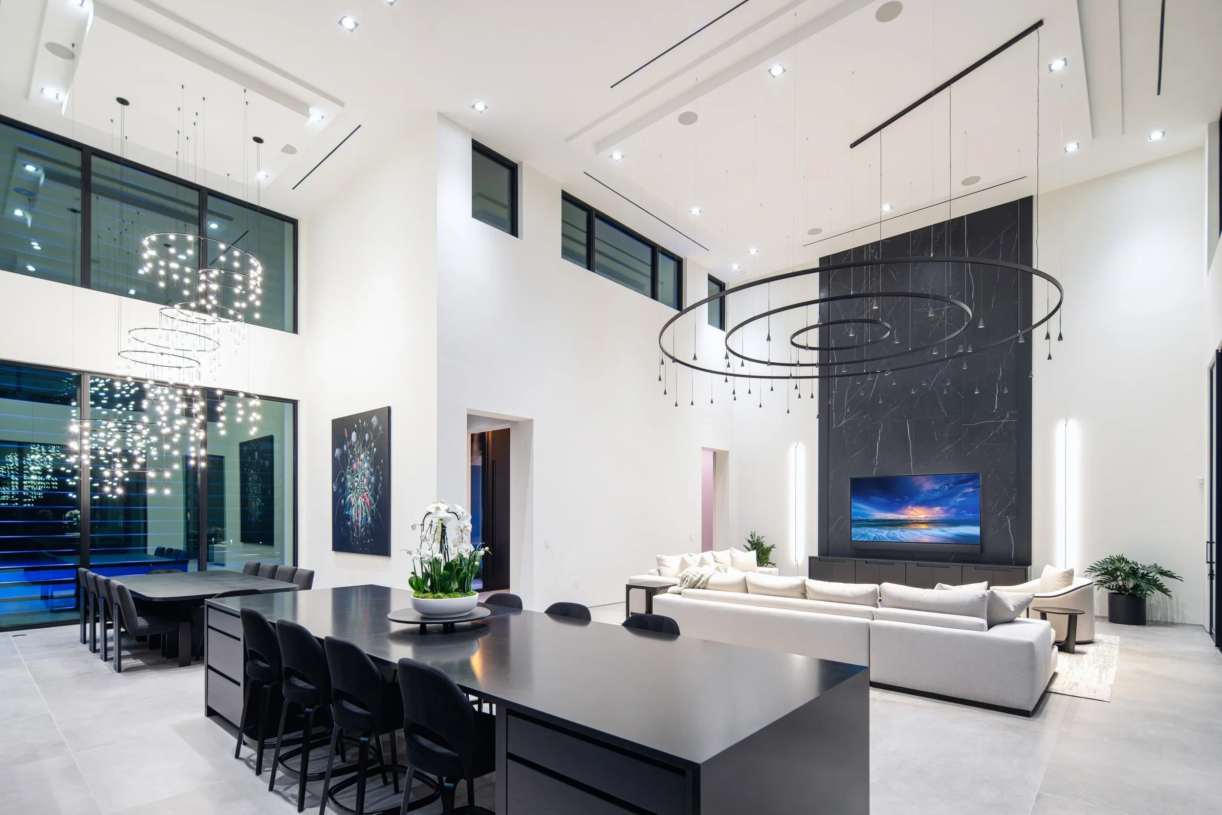 Modern living room with high ceilings, large black and white marble fireplace, wall-mounted TV, contemporary lighting, large white sectional sofa, black dining table with chairs, and large windows with city view.
