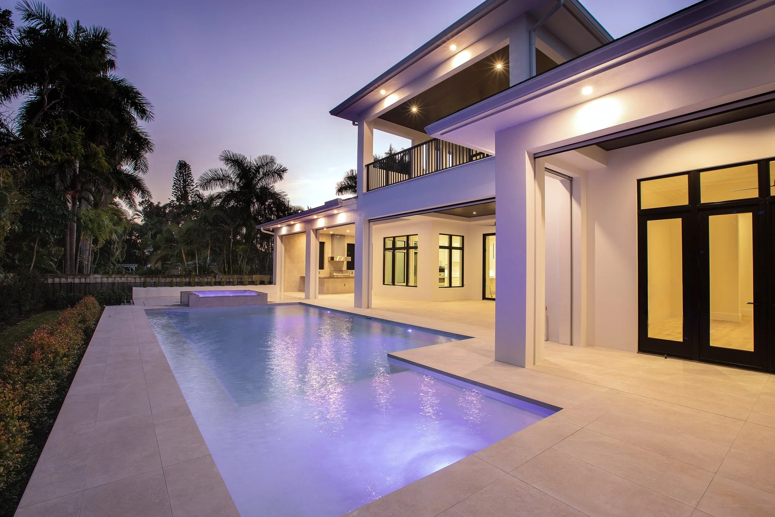 Modern house with a swimming pool in the backyard during dusk, with outdoor lighting and tropical trees in the background.