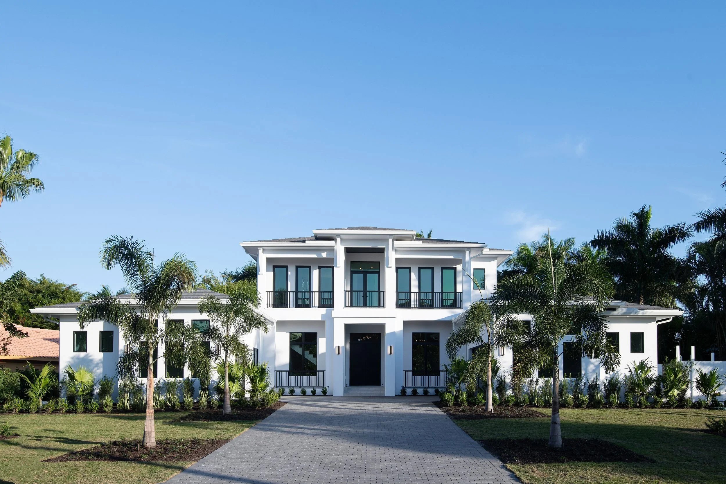 White modern two-story house with balconies, surrounded by palm trees, under a clear blue sky.