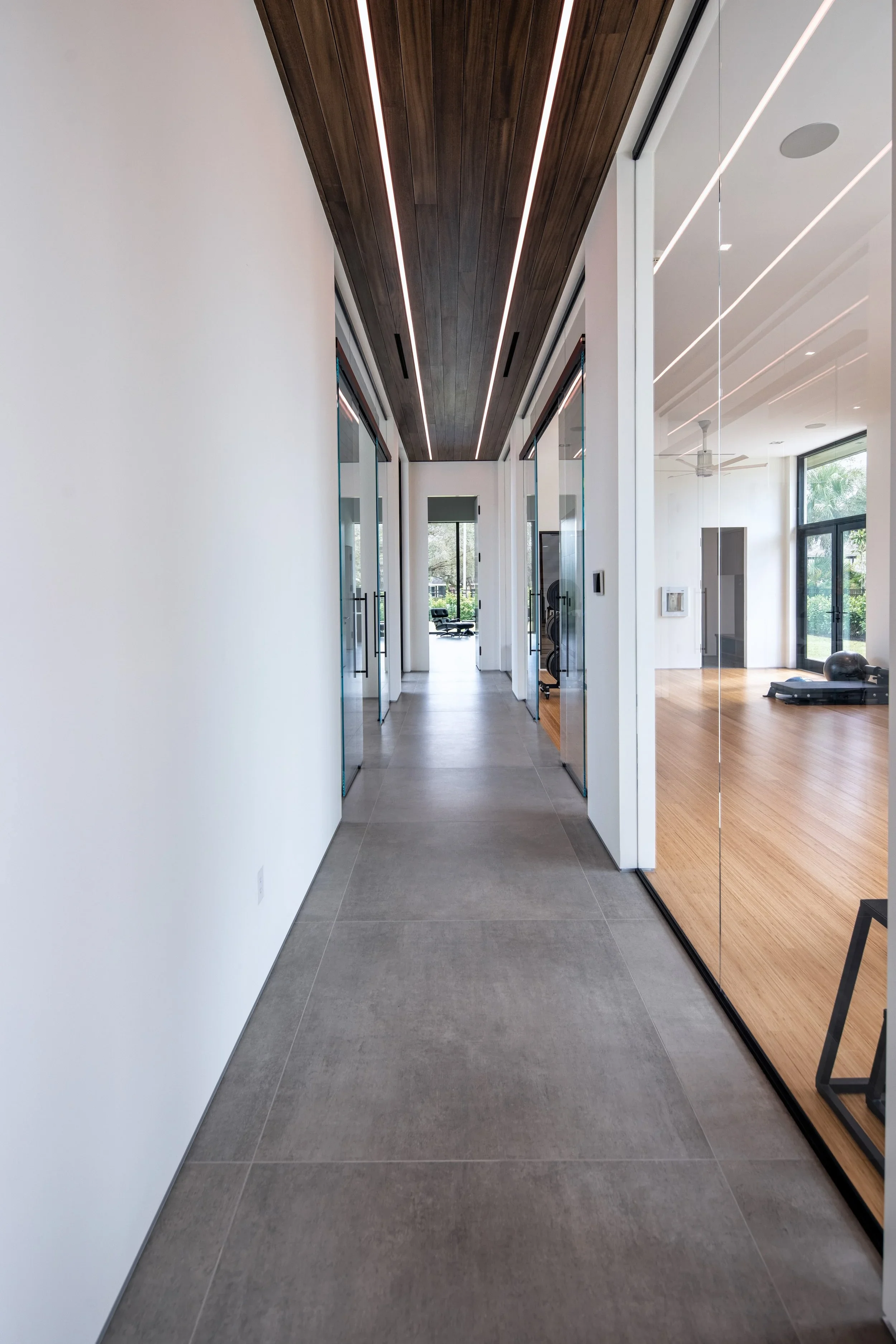 Modern hallway with grey tiled flooring, white walls, and a wooden ceiling featuring parallel LED strip lights, leading to a glass door and views of a backyard.