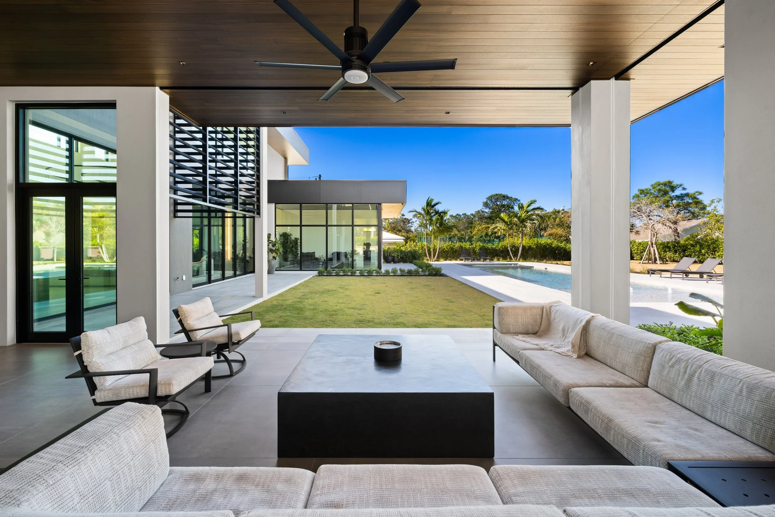 Modern outdoor patio with beige cushioned seating, a black coffee table, and ceiling fan, overlooking a grassy yard and swimming pool with palm trees under a clear blue sky.