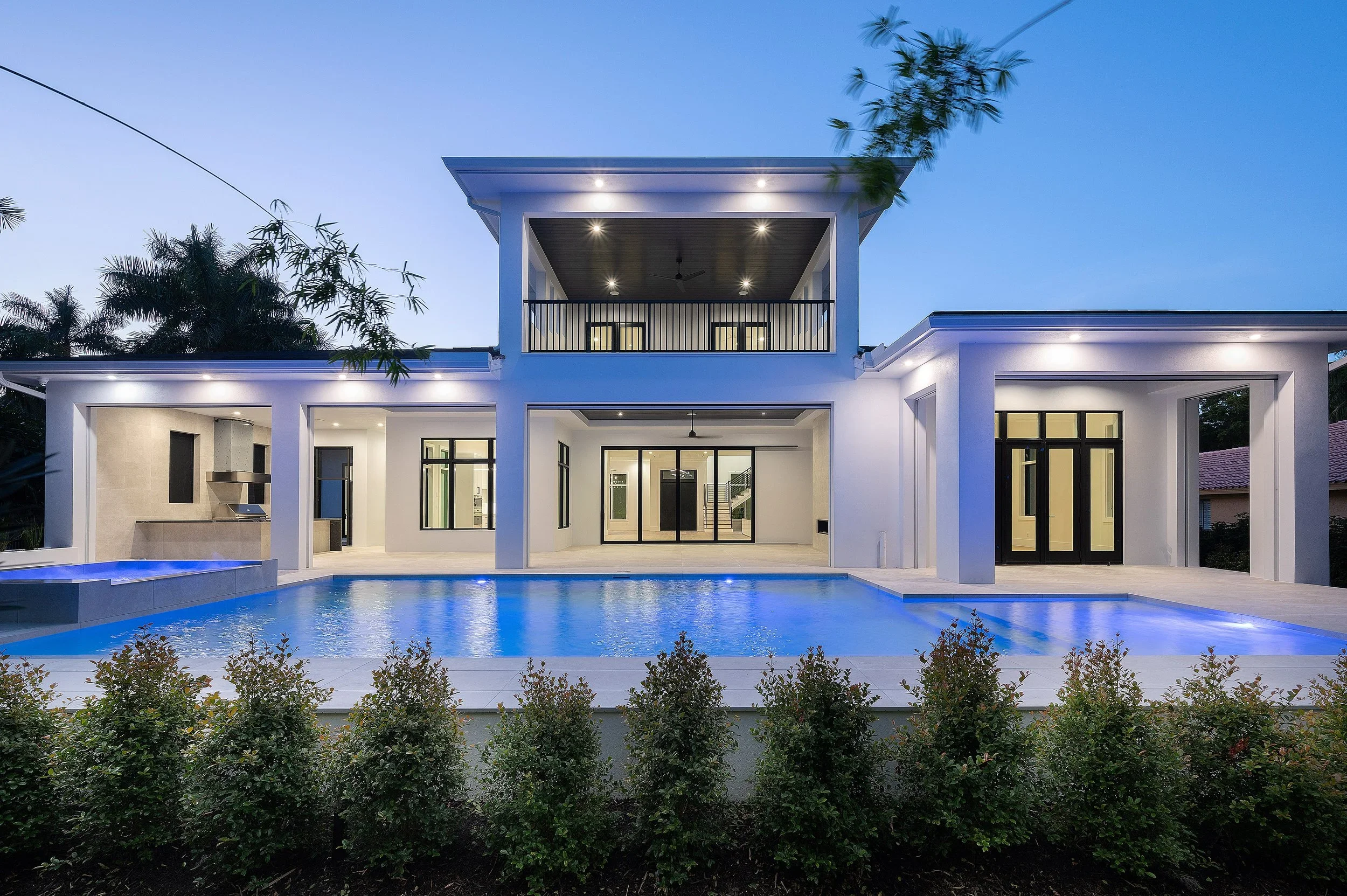 Modern two-story house with large glass sliding doors, a pool in front, outdoor lighting, and a patio area, set against a twilight sky.