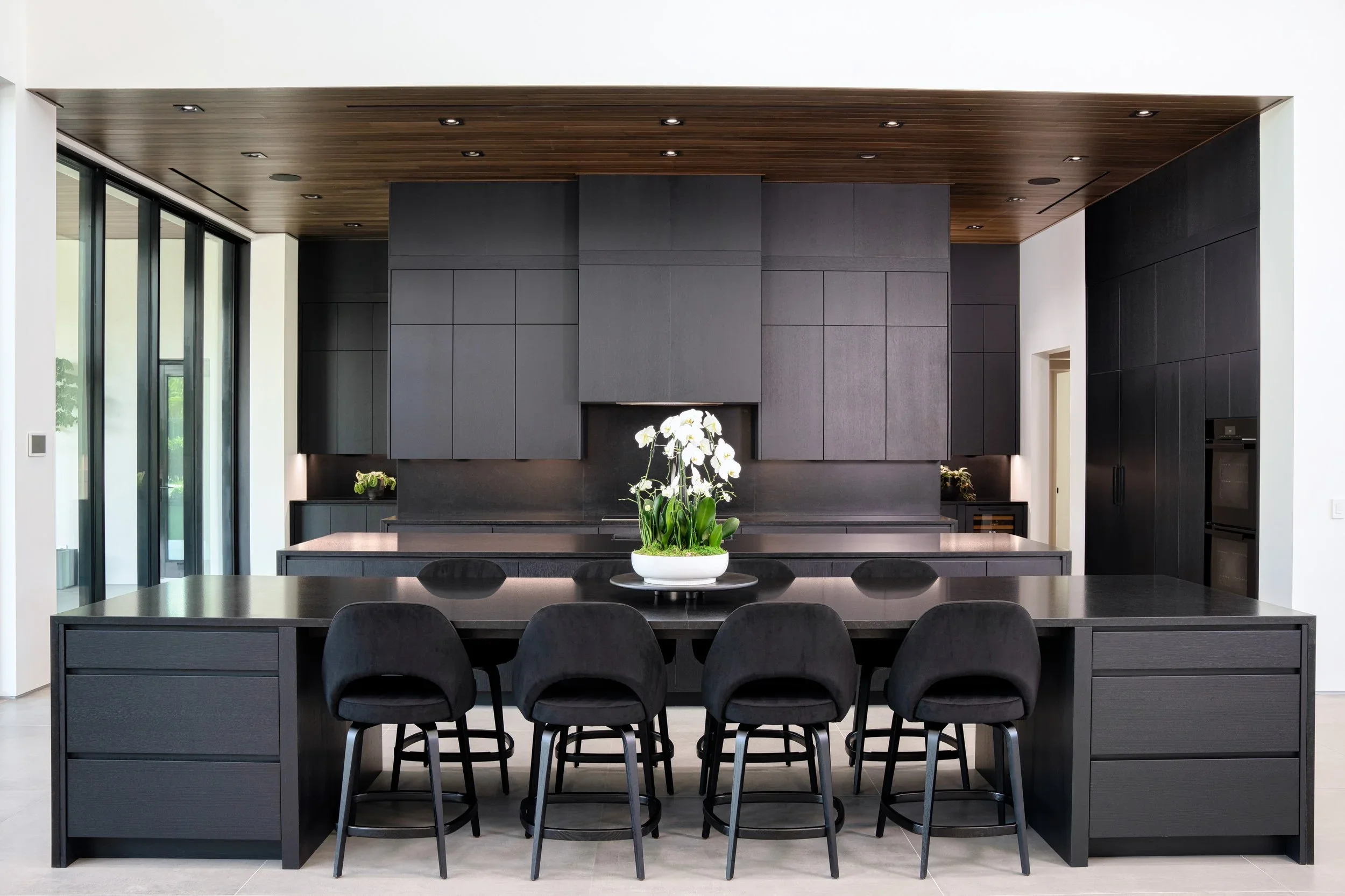 Modern kitchen with black cabinetry and black countertops, centerpiece with white orchids on island, wood ceiling, sliding glass doors on left.