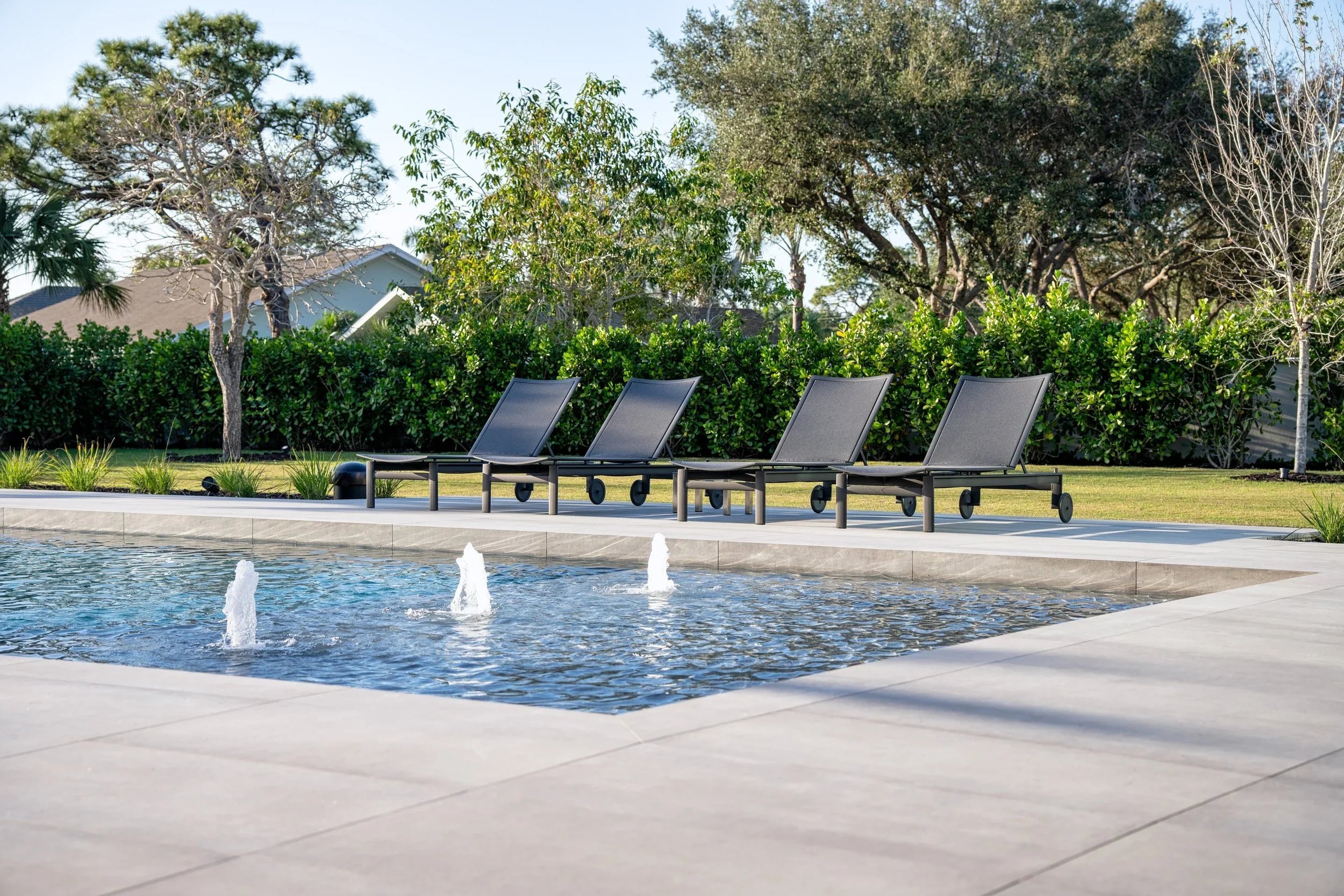 Empty poolside with four black lounge chairs, small fountains in the pool, surrounded by trees and greenery under clear sky.