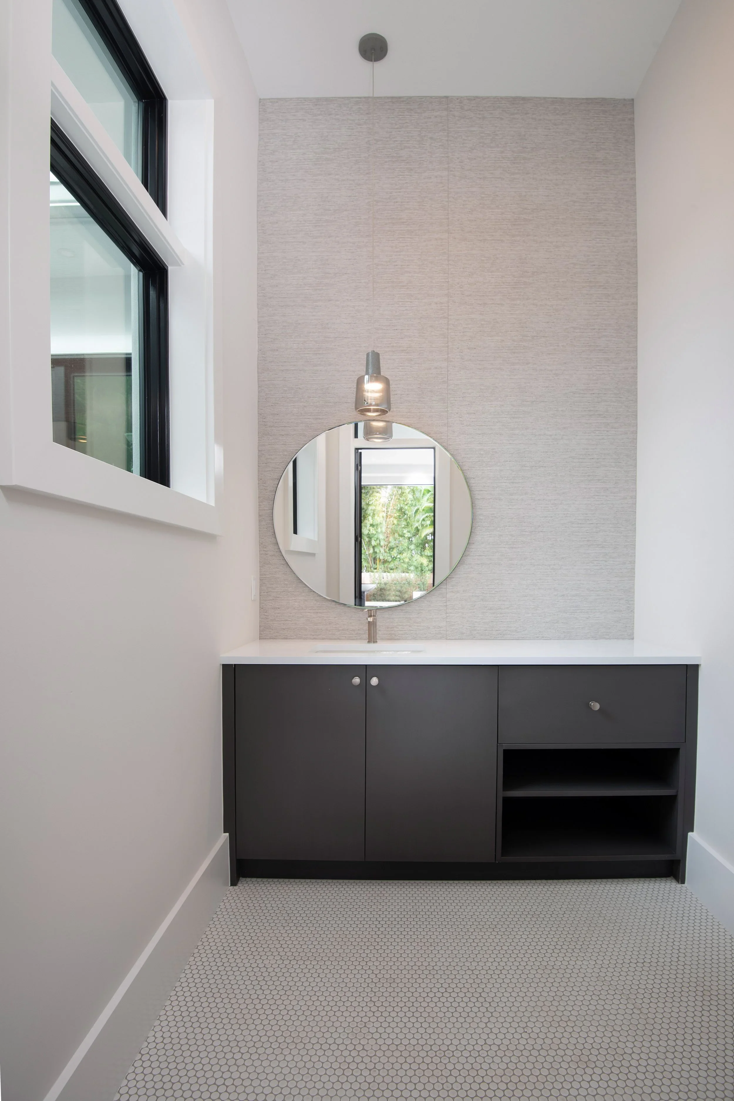 Modern bathroom vanity with a round mirror, white countertop, black cabinet, and gray textured wall behind the mirror. A window with black framing reflects greenery outside.