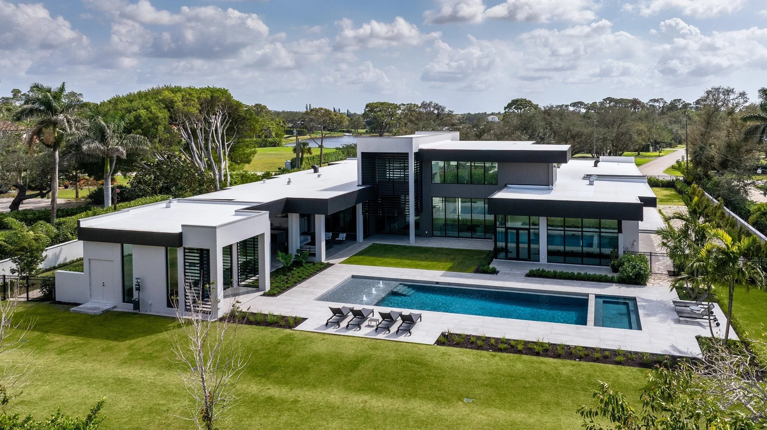 A modern house with large glass windows, a pool with lounge chairs, surrounded by a well-maintained lawn and trees under a partly cloudy sky.
