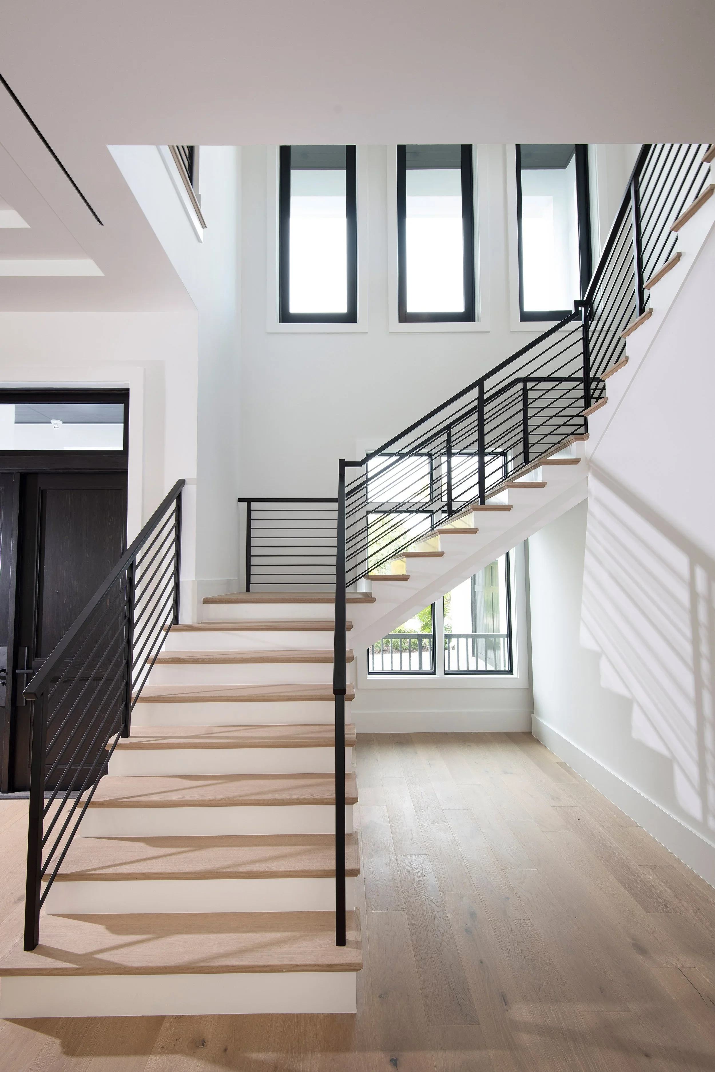 Interior of a modern home with a staircase featuring wooden steps and a black metal railing, large windows letting in natural light, and a light-colored wooden floor.