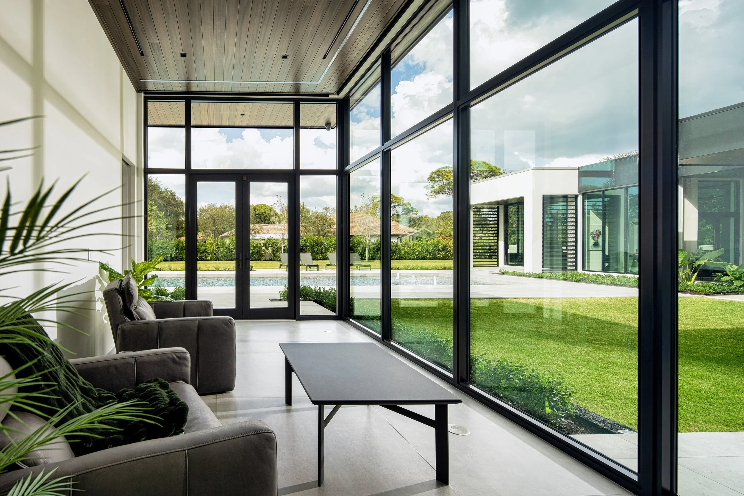 Modern living room with large glass windows looking out onto a backyard with a pool and lawn, interior includes gray sofa and black coffee table.