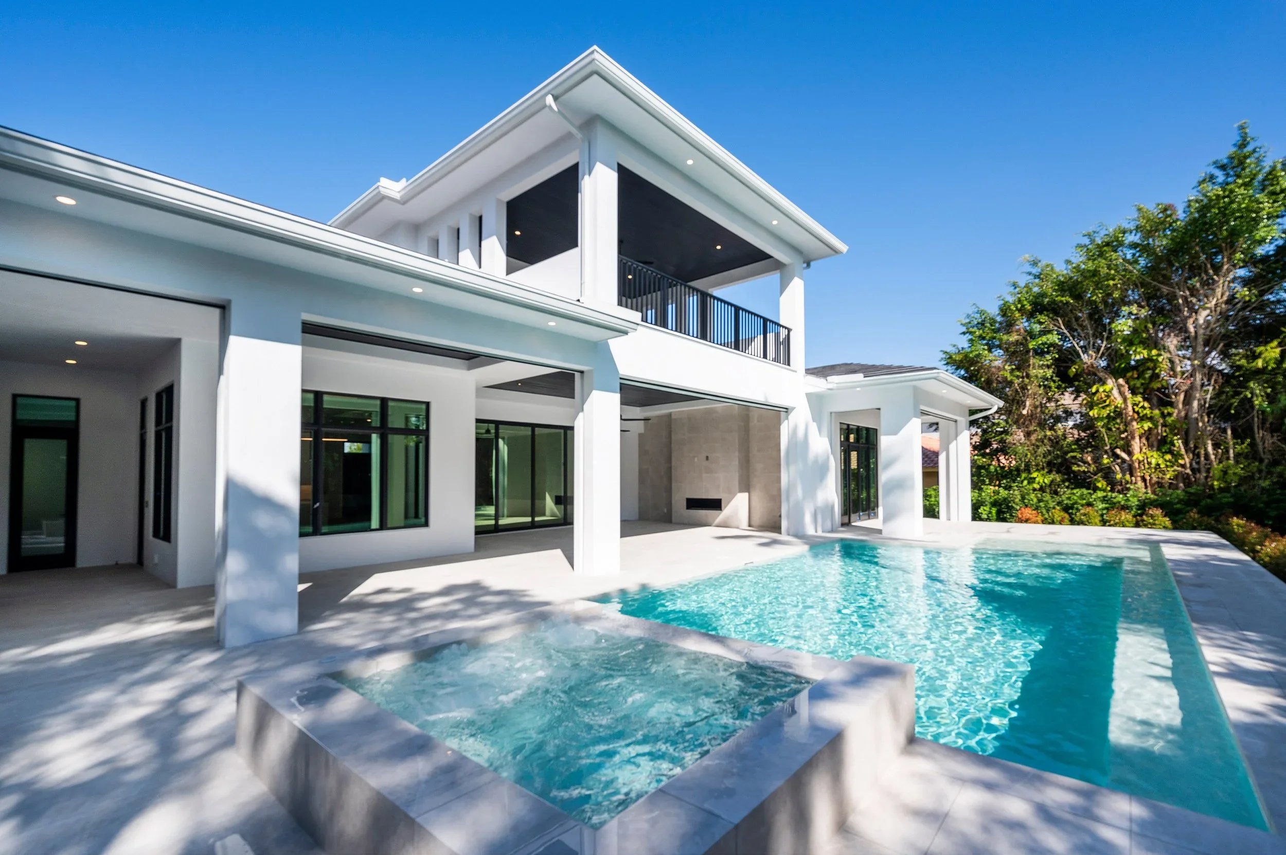 Modern white house with large outdoor pool, hot tub, and glass doors, surrounded by greenery and blue sky.