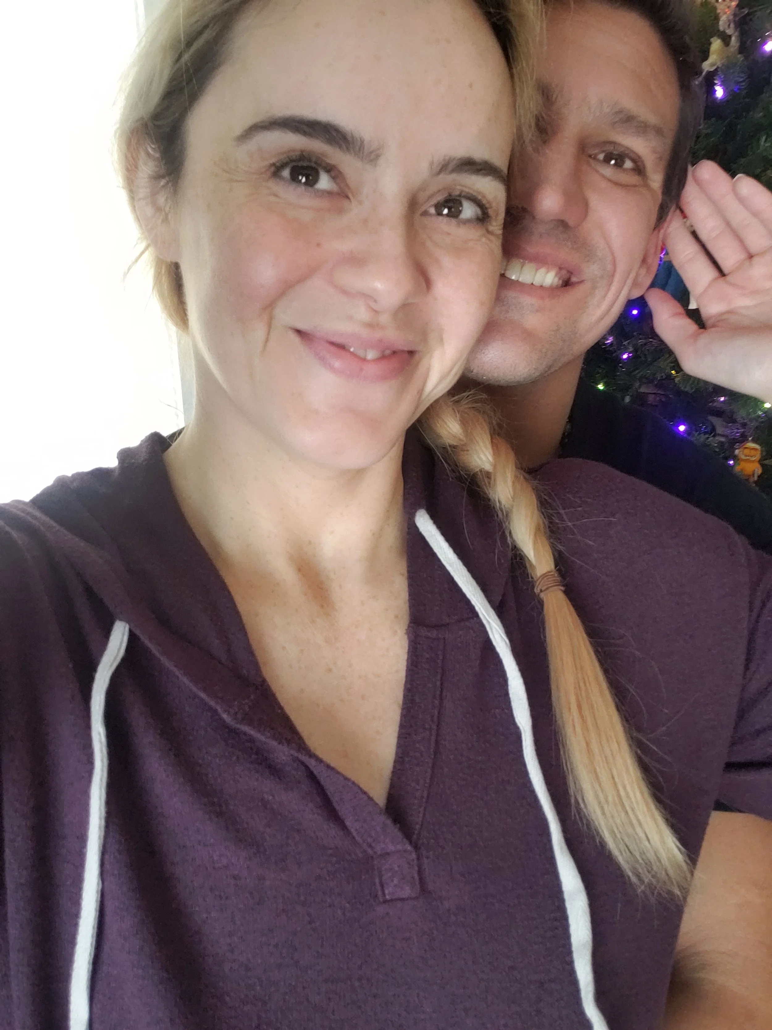 A woman and a man smiling for a selfie in front of a Christmas tree decorated with purple lights.