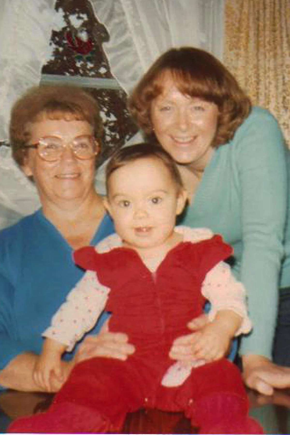 A family of three, including an elderly woman with glasses, a middle-aged woman, and a young boy, sitting together indoors in front of a Christmas tree decorated with ornaments and a star on top. The boy is sitting on the elderly woman's lap, and everyone is smiling.