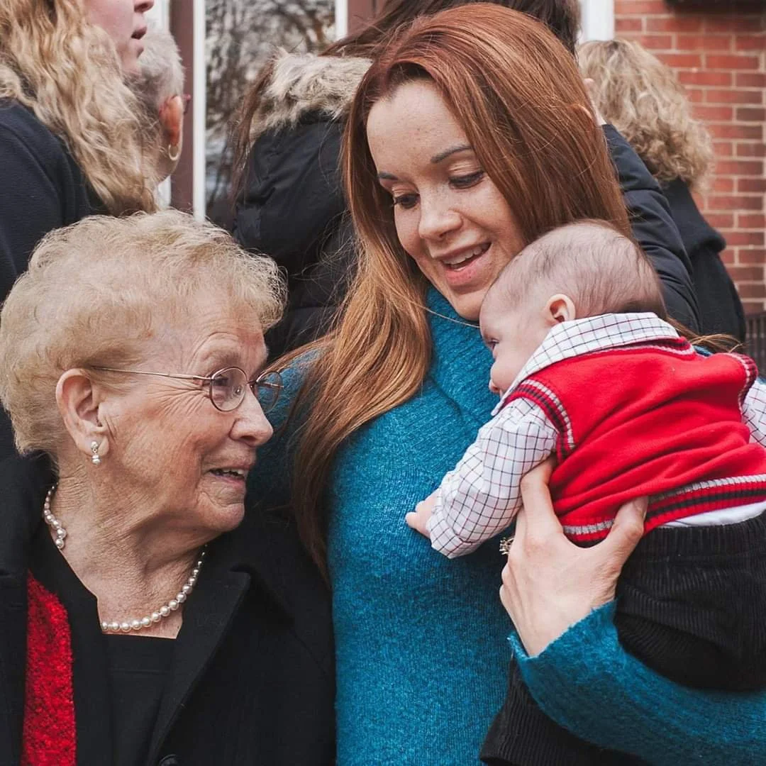 An elderly woman, a young woman, and a baby engaged in a warm, smiling interaction outdoors against a brick building. The elderly woman is wearing glasses and pearl earrings, the young woman has long red hair, and the baby is dressed in a red vest and checked shirt.
