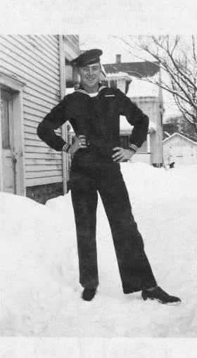 A person dressed in a navy sailor uniform standing outdoors in snowy weather, with houses and trees in the background.