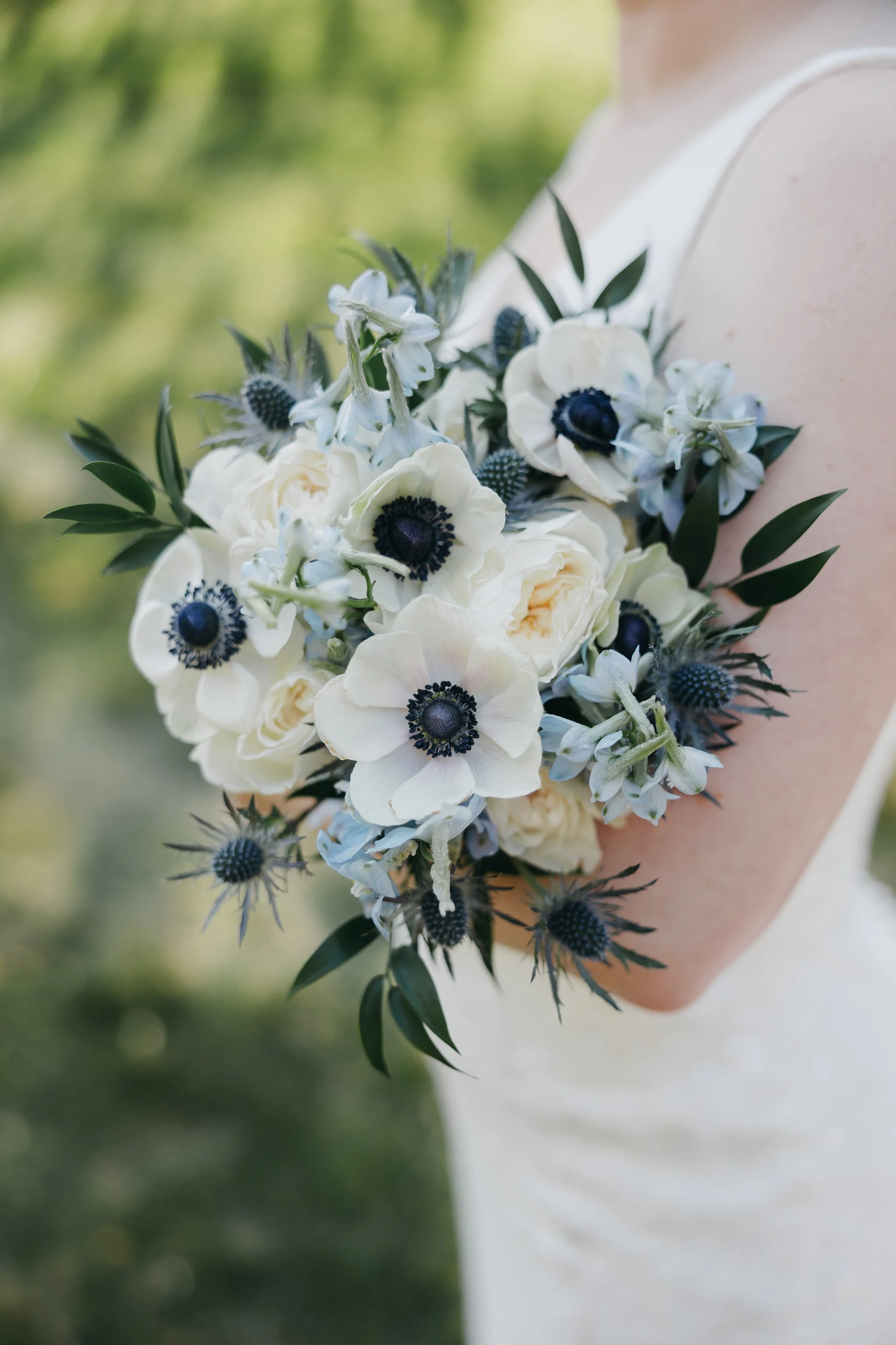 Bride holding a bouquet of white and blue flowers, including anemones, roses, and greenery, outdoors with a blurred green background.
