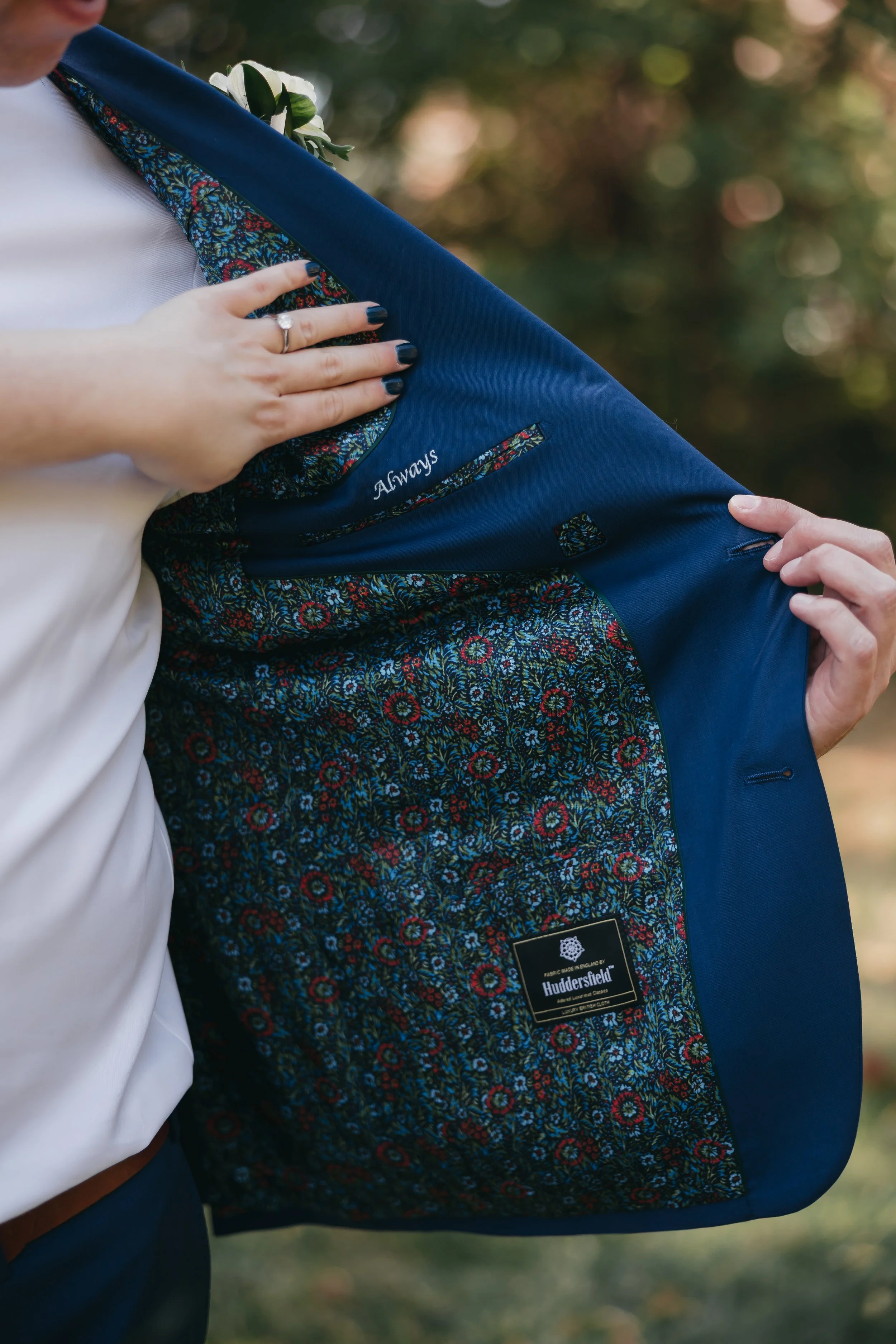 Close-up of a person holding open a navy blue blazer, revealing a floral patterned lining and a label that reads "Huddersfield." The person is wearing a white shirt and has a ring on their finger, with a blurred outdoor background.