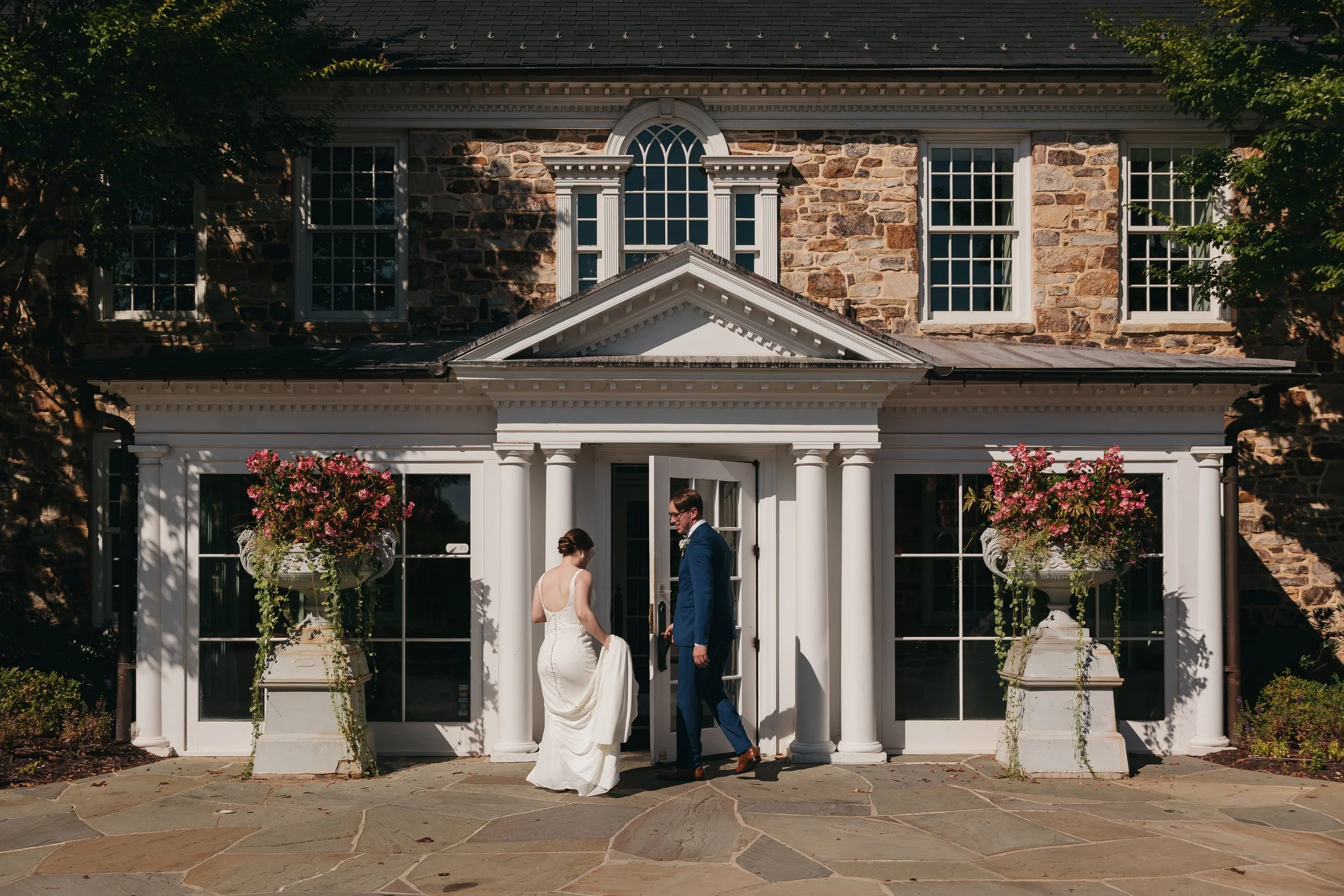 A bride and groom standing outside a historic stone and white wooden house with columns, large windows, and flower pots on pedestals.