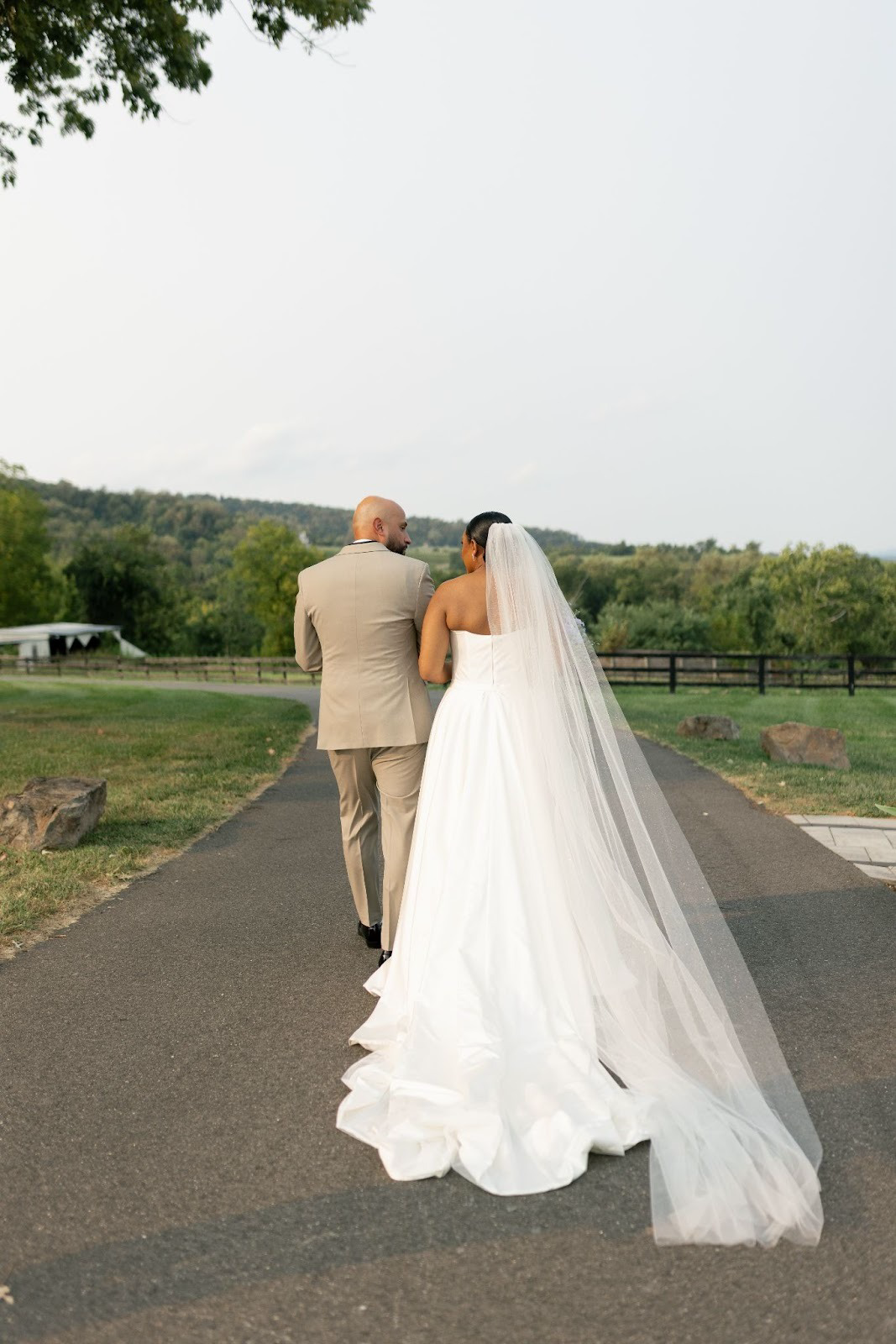 A bride and groom walking on a paved path outdoors with a scenic green landscape and distant hills, the bride in a white wedding dress with a veil, and the groom in a beige suit.