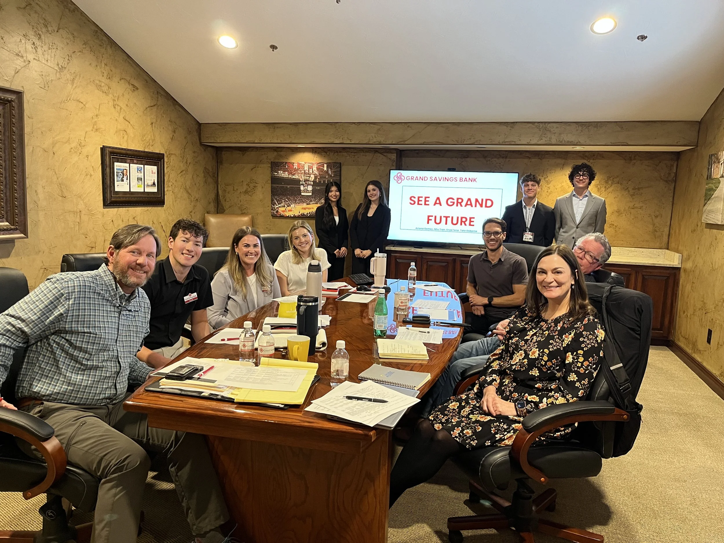 A group of people sitting around a conference table in a meeting room, smiling for a photo with a large screen displaying 'SEE A GRAND FUTURE' and 'Grand Savings Bank' in the background.
