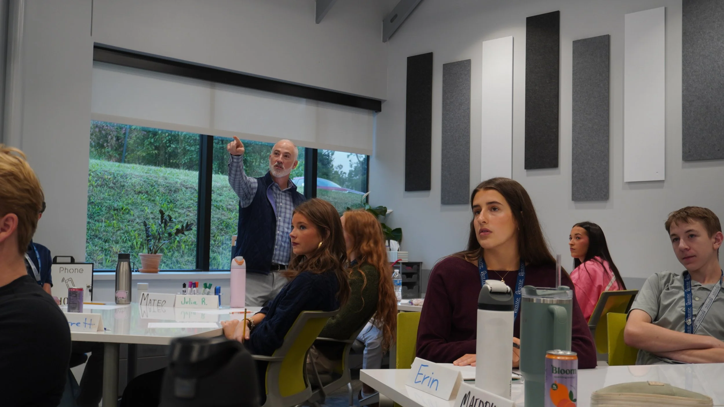 A classroom or workshop setting with students sitting at tables, attentively listening to an older male instructor who is standing near a window and pointing upwards.