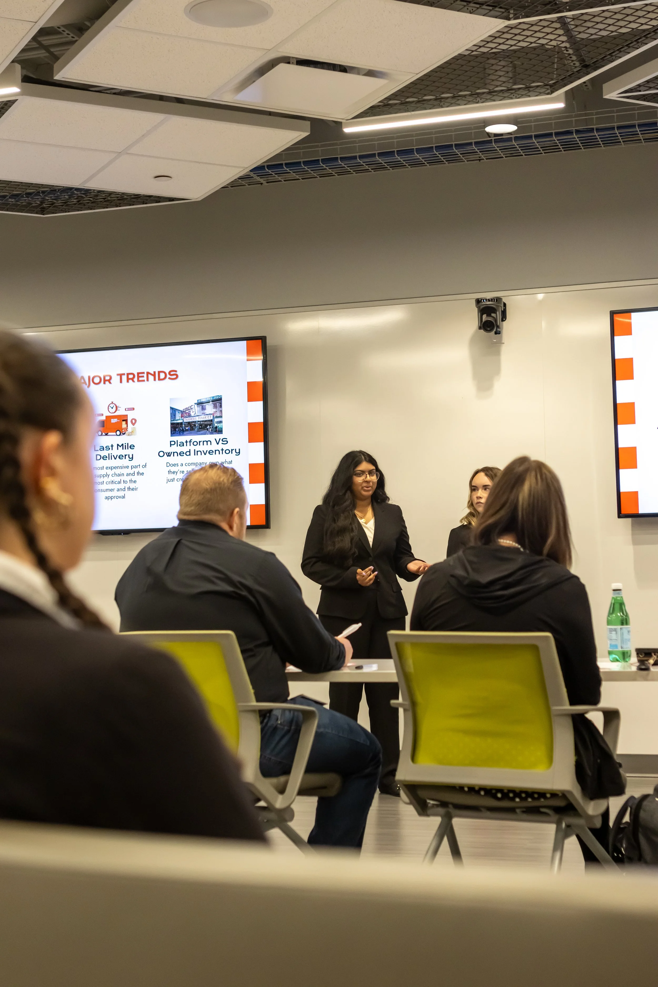 A woman presenting during a business meeting or conference, with attendees seated and a presentation slide titled 'Major Trends' displayed on a screen behind her.