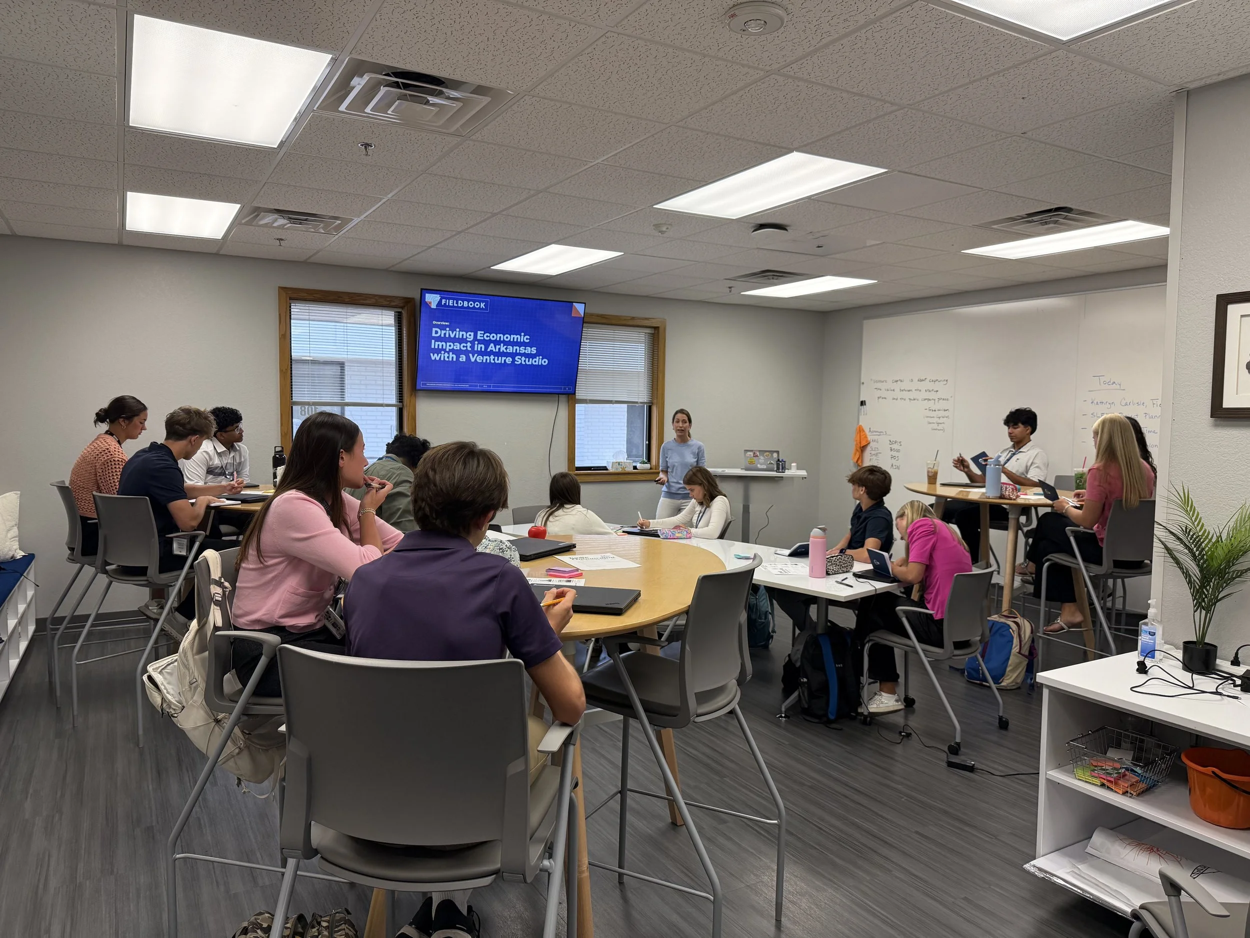 A classroom with students seated around a large oval table, a teacher standing at the front near a whiteboard, and a large screen displaying a presentation titled 'Driving Economic Impact in Arkansas with a Venture Studio'.