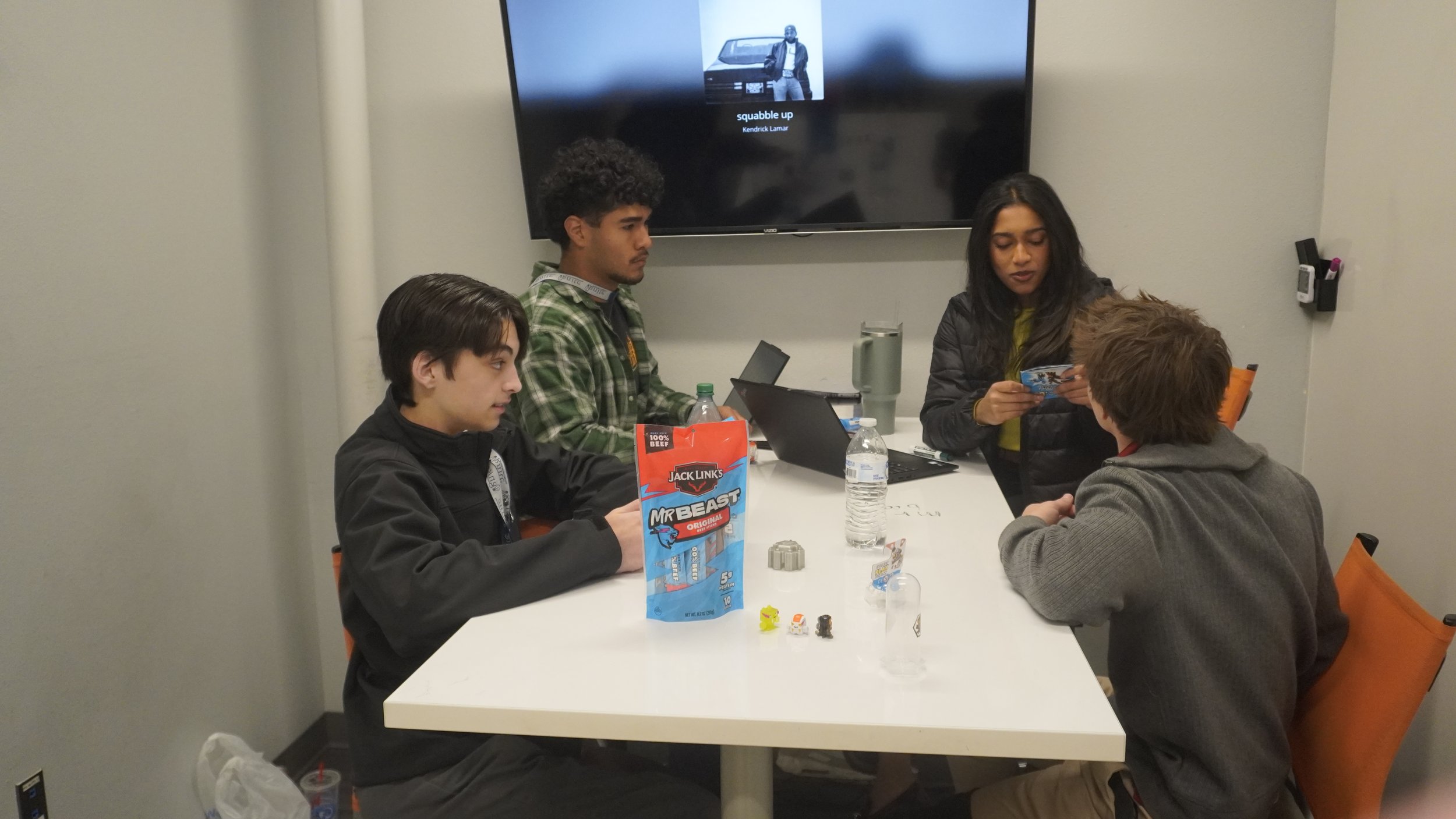 Four young people sitting around a white table in a room with gray walls, engaging in a tabletop game. There are various game pieces, water bottles, and laptops on the table. A large screen on the wall displays song information.