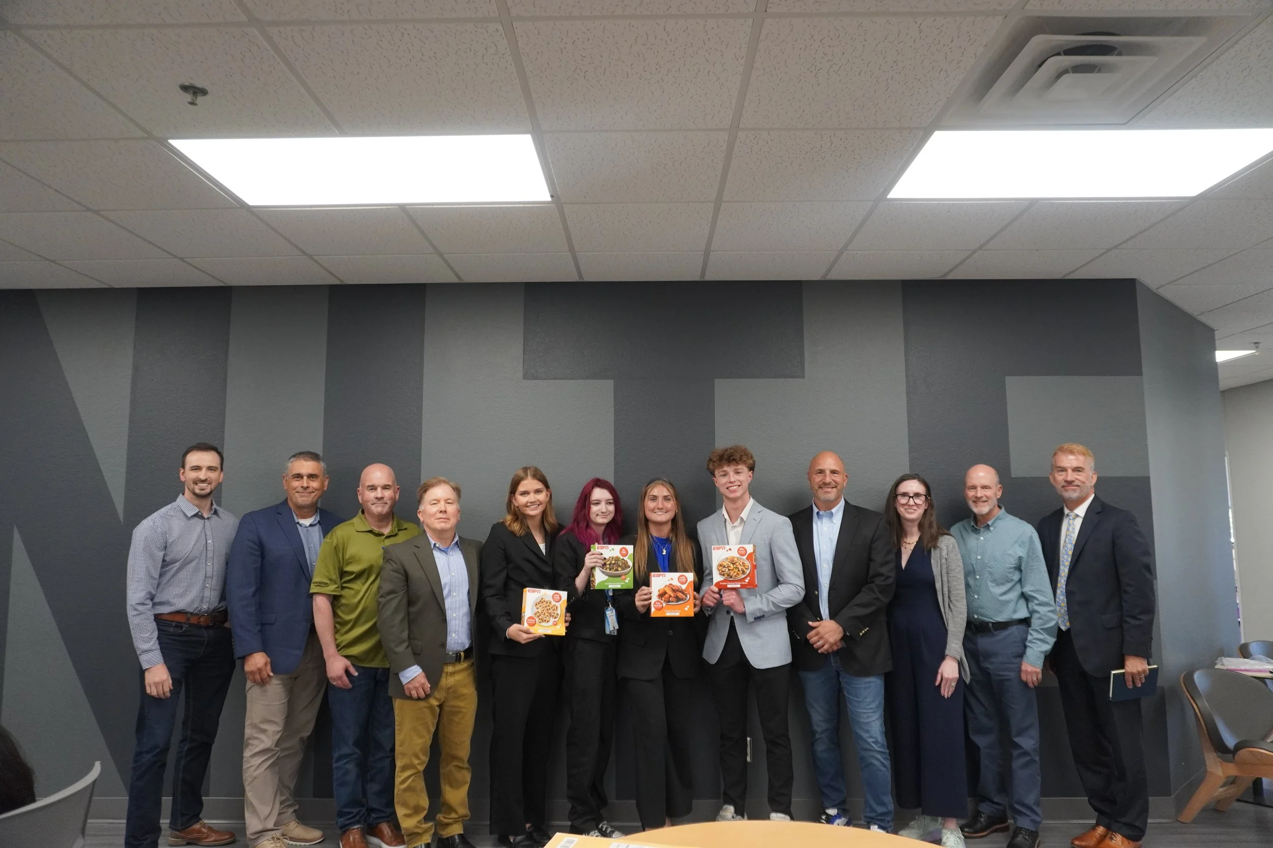 Group of fifteen people standing in a row in an office space, smiling, with some holding food product boxes.
