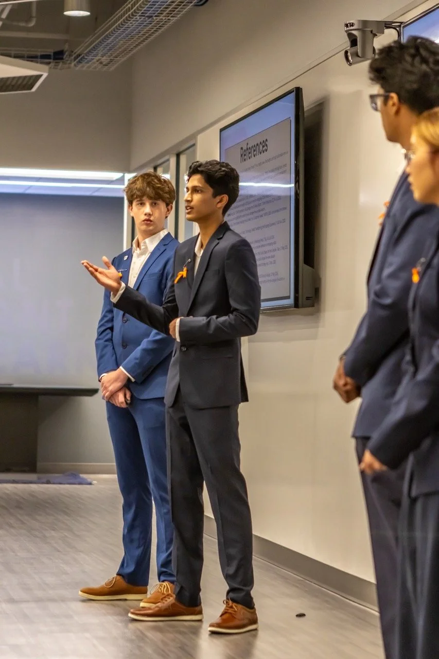 A group of young professionals giving a presentation in a conference room, with a large screen displaying a slide titled 'References'. The man in front is speaking and gesturing with his hand, wearing a dark suit, and others are listening attentively