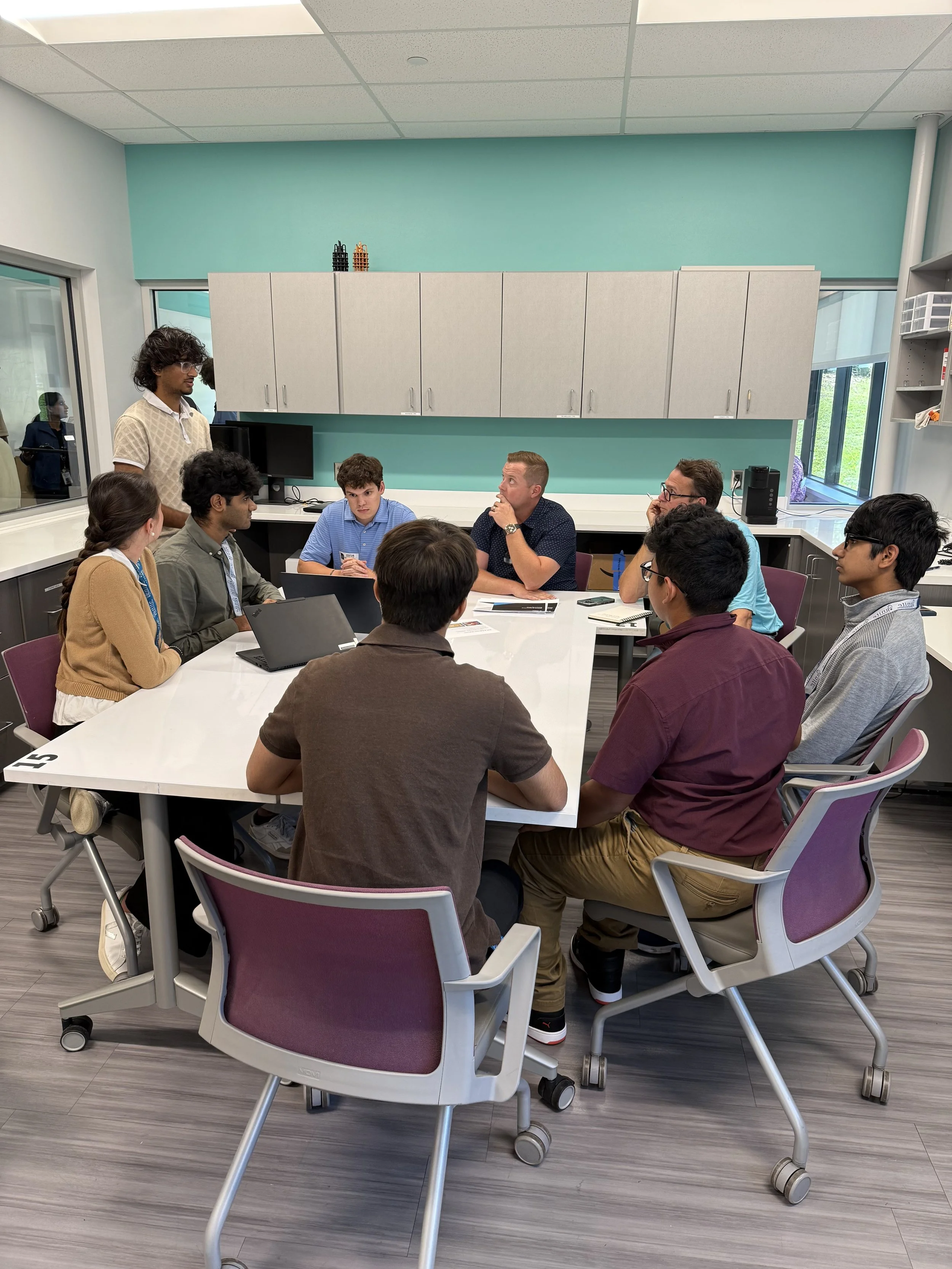 A diverse group of nine young professionals gathered around a white conference table in a modern meeting room, with one person standing and speaking while others listen. The room has teal and white walls, cabinets, and windows allowing natural light.